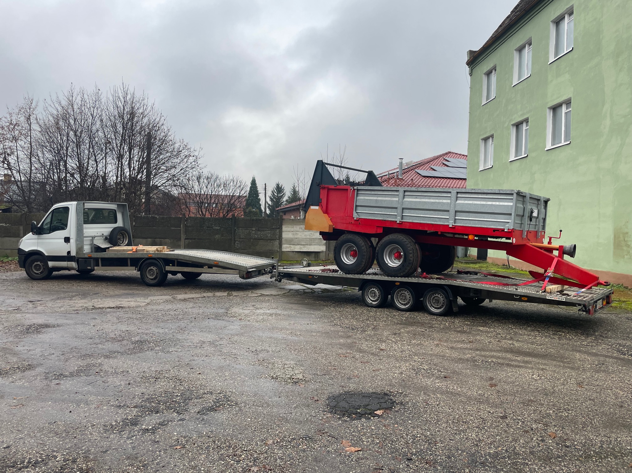 Biała laweta z platformą przewozi czerwoną przyczepę rolniczą na tle budynku z zieloną elewacją i pochmurnego nieba. Widok z boku na placu.