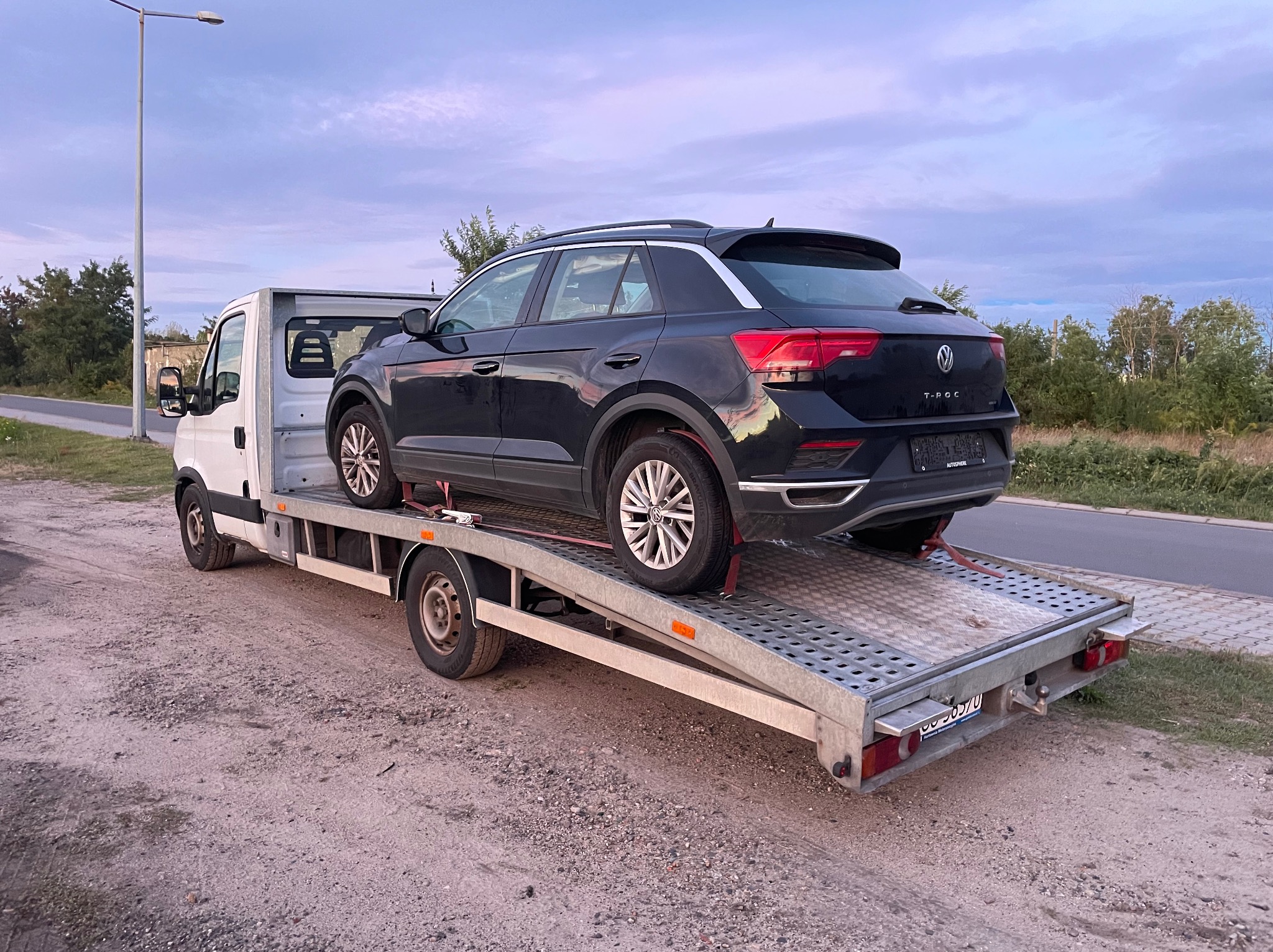 Czarny Volkswagen T-Roc na lawecie z ażurową platformą, zabezpieczony czerwonymi pasami. Widok z tyłu na szarym tle nieba i drzew.