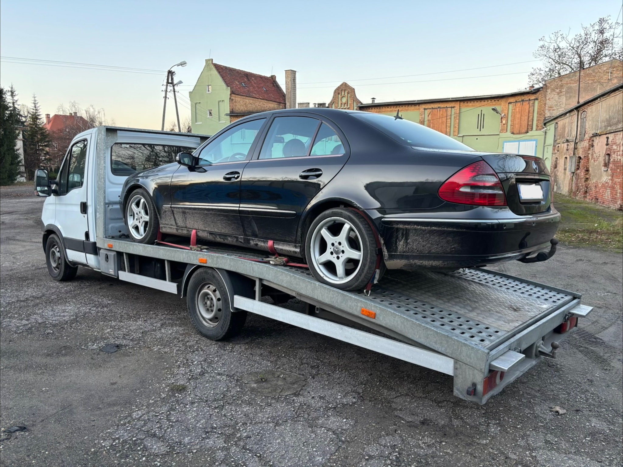 Czarny Mercedes na srebrnej lawecie z najazdami, zabezpieczony czerwonymi pasami. W tle budynki i drzewa. Usługa transportu pojazdów.