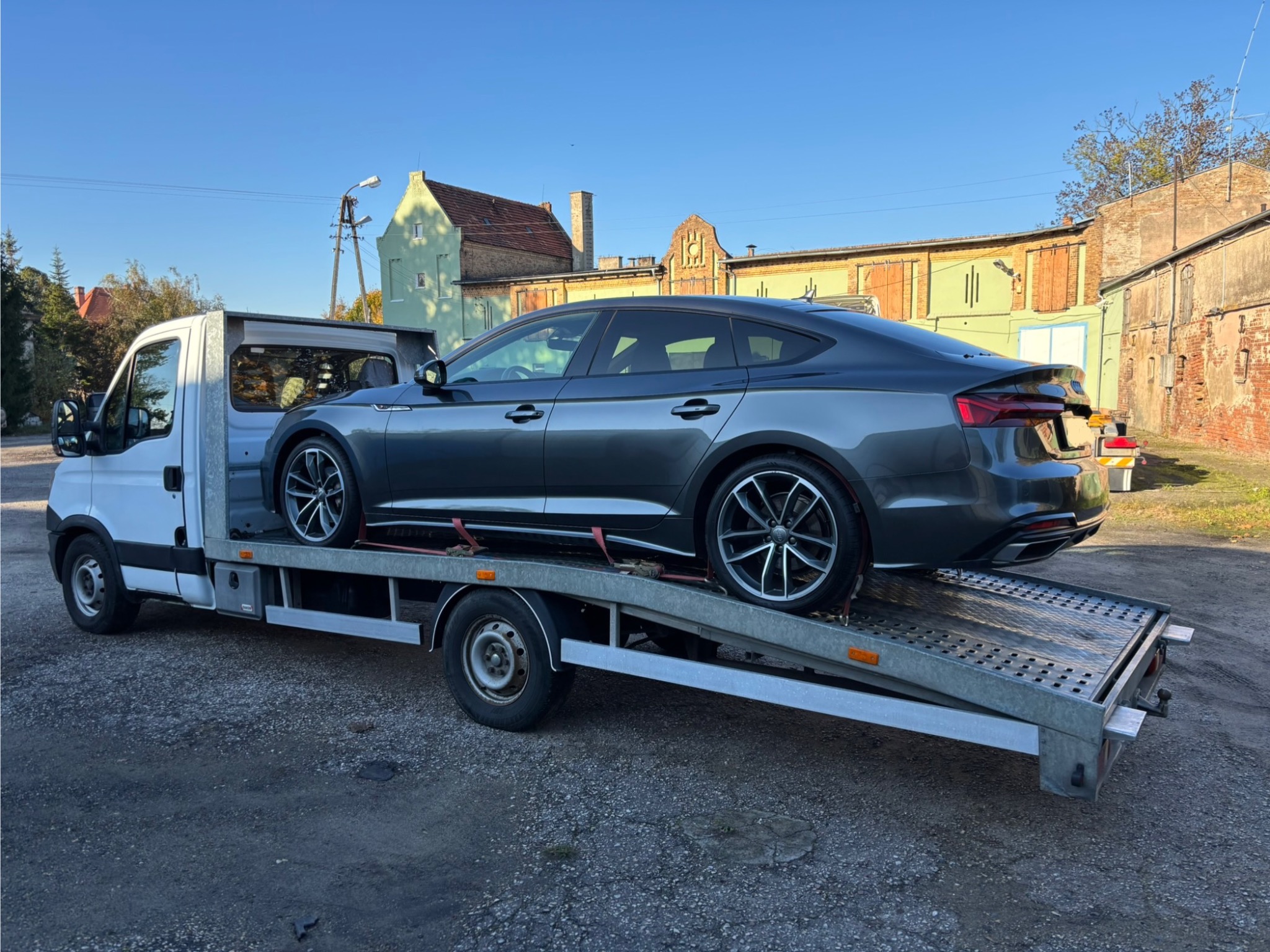 Szary samochód osobowy marki Audi A5 na platformie lawety, zabezpieczony czerwonymi pasami transportowymi. W tle budynki i błękitne niebo.