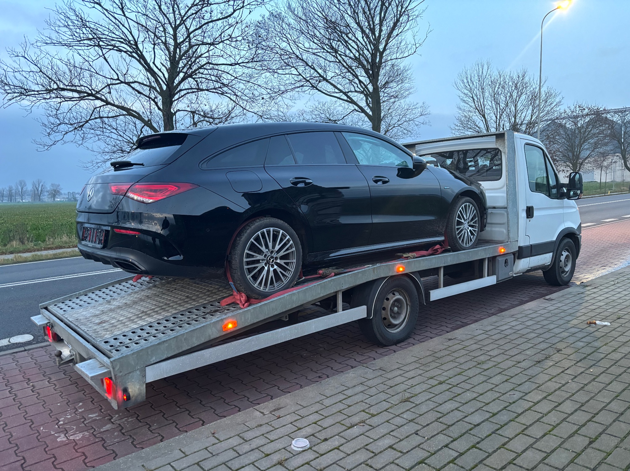 Czarny Mercedes CLA kombi zabezpieczony pasami na platformie lawety, widok z tyłu, szara kostka brukowa i drzewa w tle.