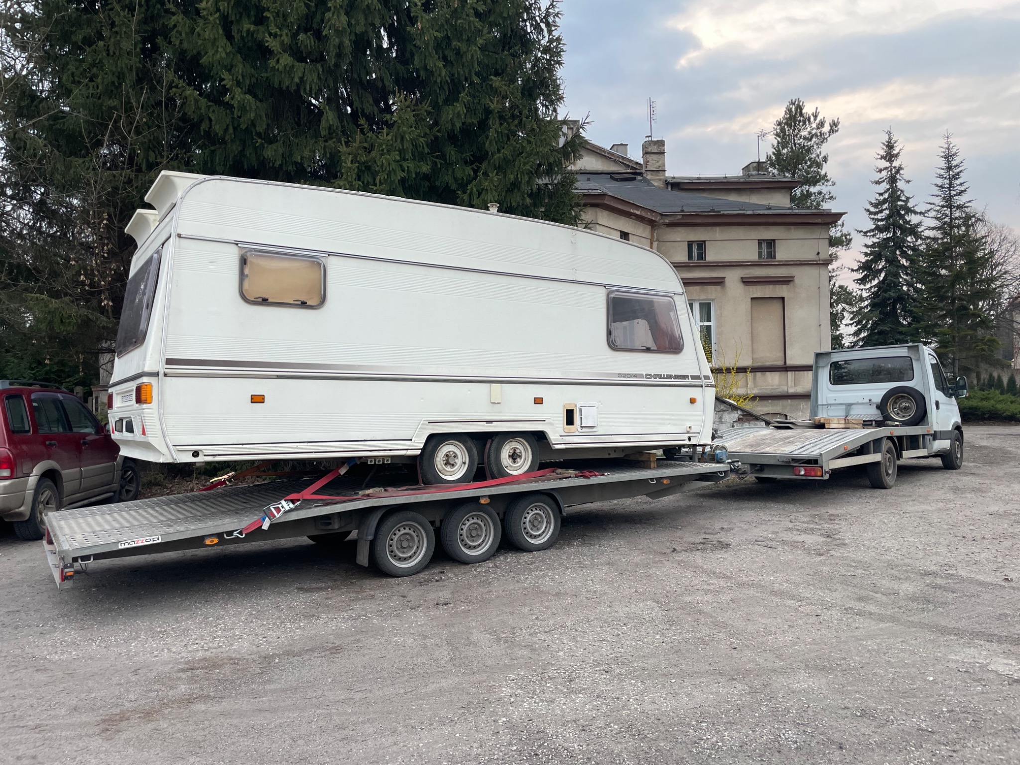 Laweta przewozi białą przyczepę kempingową na platformie. Czerwone pasy zabezpieczają ładunek. W tle budynek i drzewa. Pochmurne niebo.