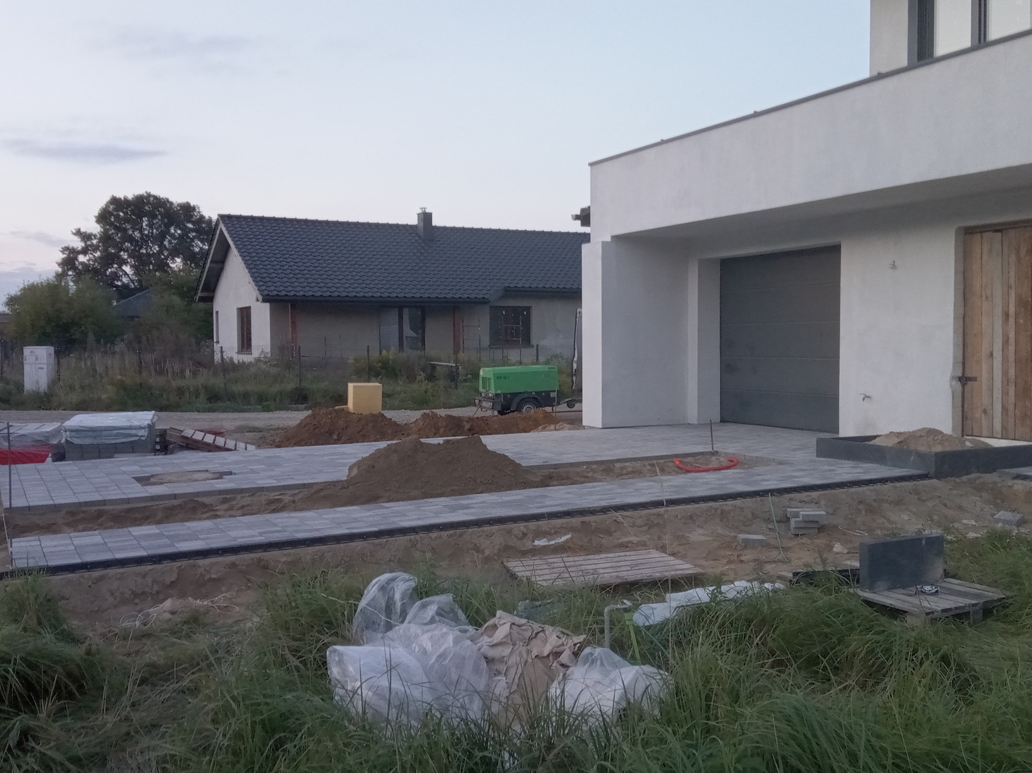 W trakcie budowy podjazdu z szarej kostki brukowej przed nowoczesnym domem z garażem, widoczne narzędzia i materiały budowlane, w tle drugi budynek mieszkalny.