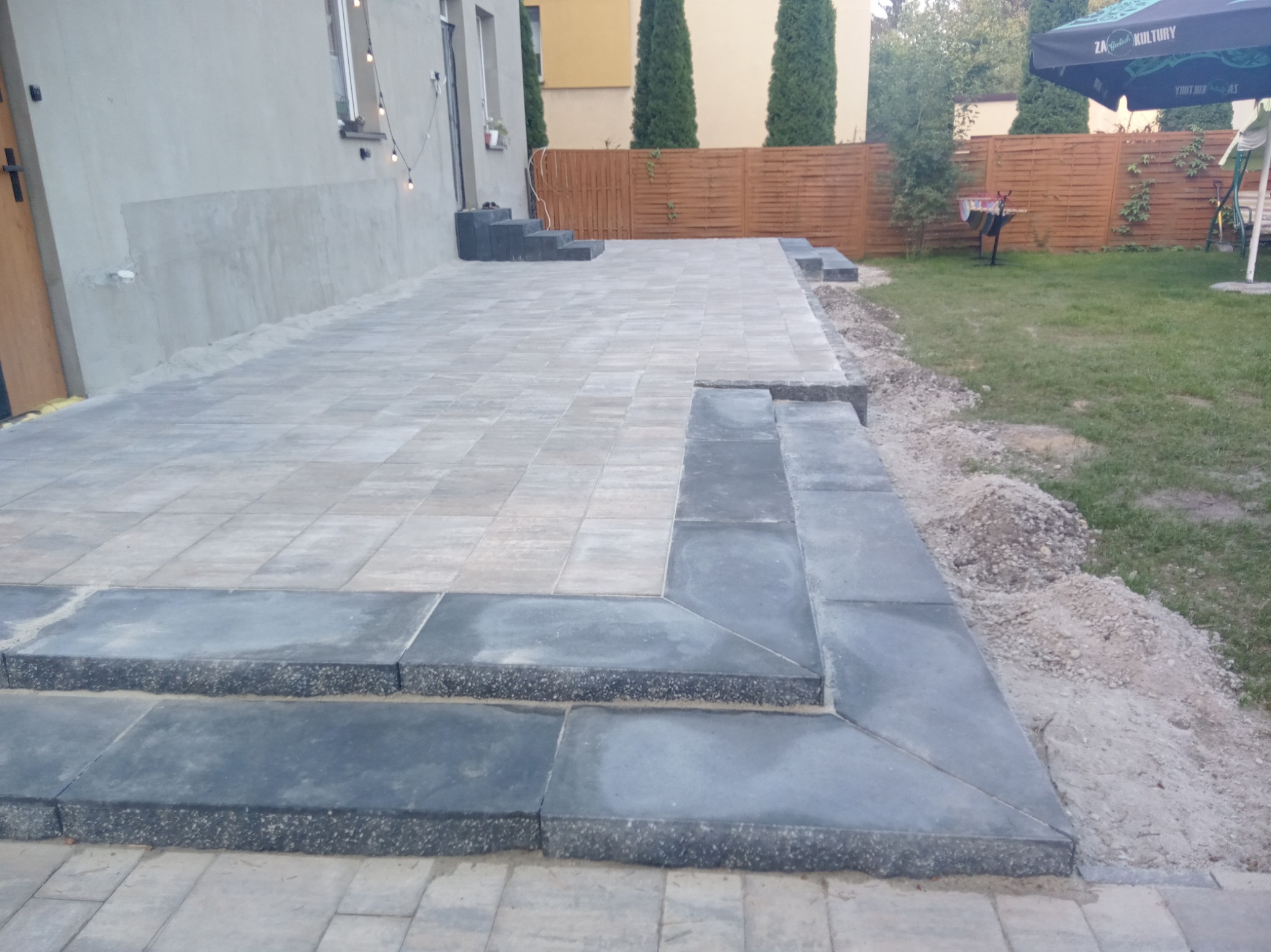 Nowo ułożony taras z kostki brukowej w odcieniach szarości, z niskimi schodami, widok na budynek z oknami i drewniany płot w tle.