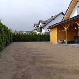 Ogrody Perfecto - Przygotowany teren pod trawnik, wyrównana ziemia, widoczne ślady po grabiach, obrzeża trawnikowe, rząd tuj na granicy działki, w tle budynki mieszkalne i drewniana altana z oświetleniem.