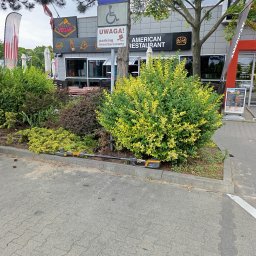 Ogrody Perfecto - Przycięte krzewy przed restauracją z narzędziem do pielęgnacji. Krzewy są zielone i żółte, a w tle widoczny jest budynek restauracji z szyldami.