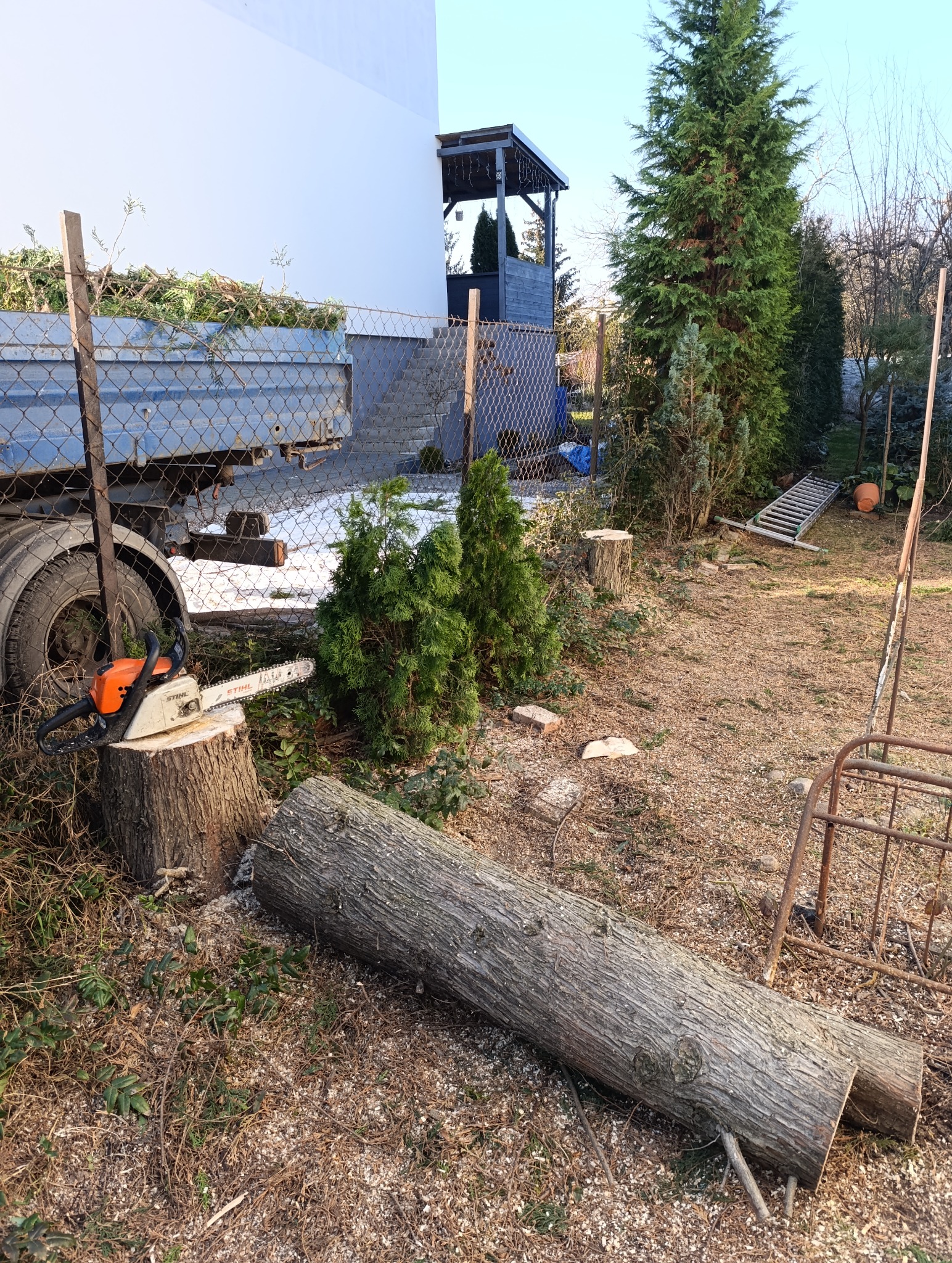 Przyczepa wypełniona gałęziami, obok leży ścięty pień, a na nim spalinowa piła łańcuchowa Stihl, w tle widoczne drzewa i elementy architektury ogrodowej.
