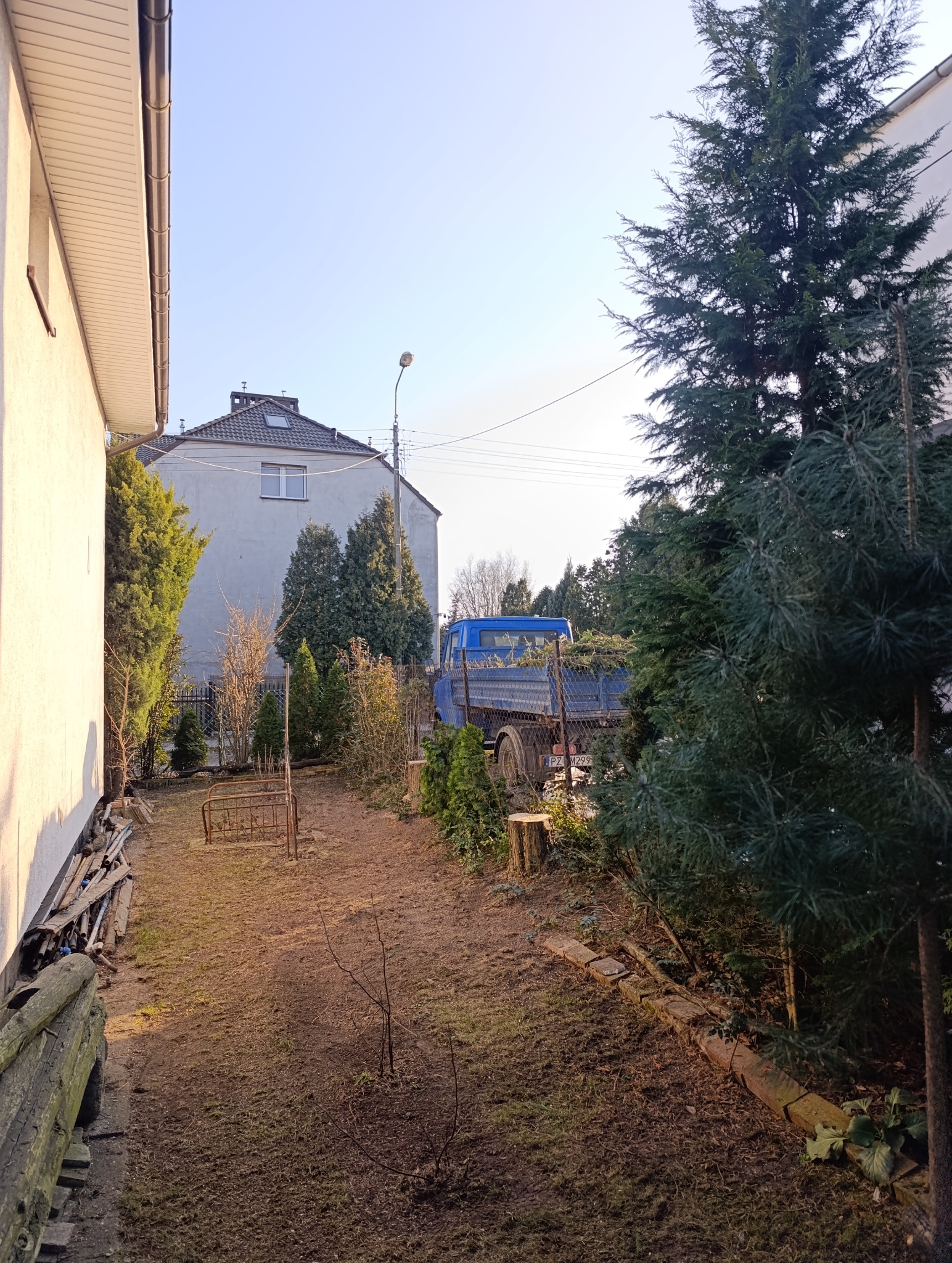 Przygotowanie terenu pod ogród w Borówcu: widok na ziemię, ścięte pnie drzew i niebieską ciężarówkę za ogrodzeniem, wypełnioną gałęziami i resztkami roślinnymi.