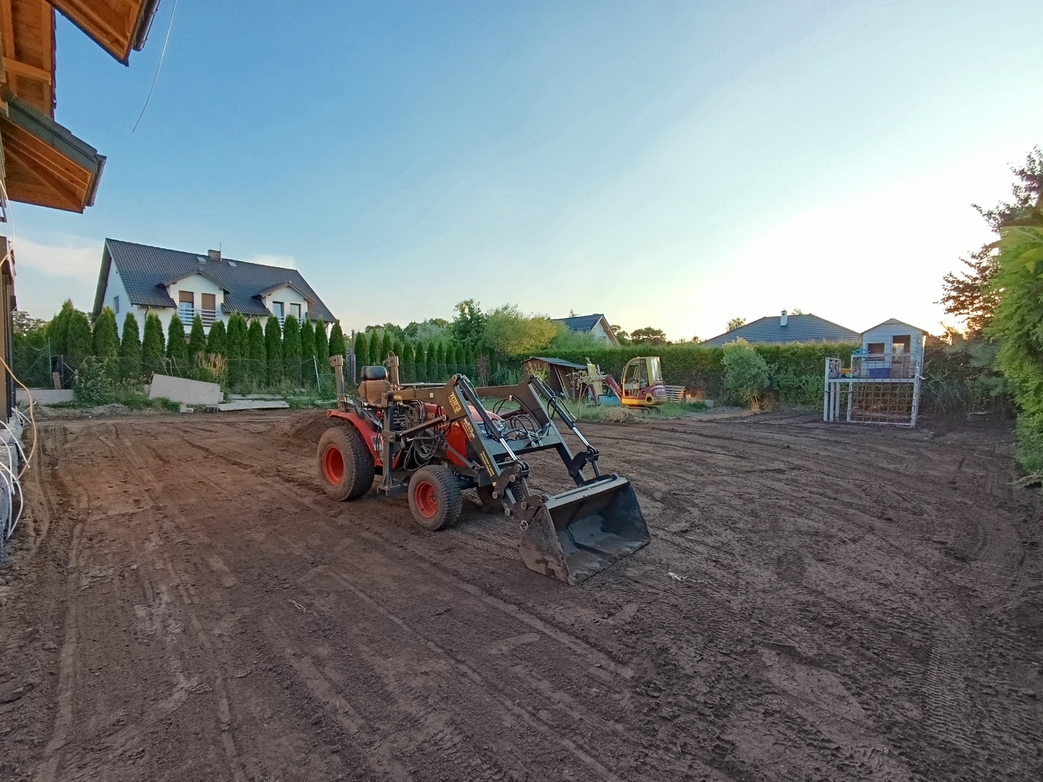 Teren przygotowywany pod ogród z użyciem małej ładowarki z czerwoną kabiną, widoczne ślady opon na ubitej ziemi, w tle dom jednorodzinny z ciemnym dachem.