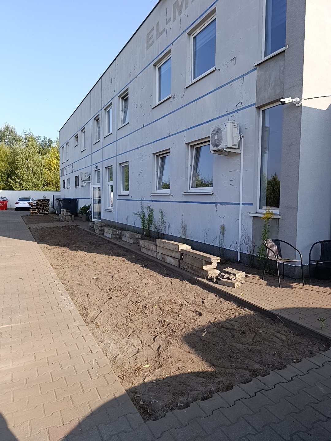 Przygotowany teren pod przyszły trawnik przed budynkiem biurowym z widocznymi paletami i krzesłami w tle.