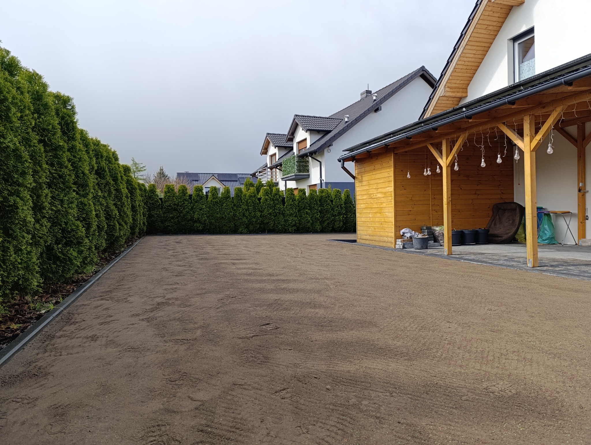 Przygotowany teren pod trawnik, wyrównana ziemia, widoczne ślady po grabiach, obrzeża trawnikowe, rząd tuj na granicy działki, w tle budynki mieszkalne i drewniana altana z oświetleniem.