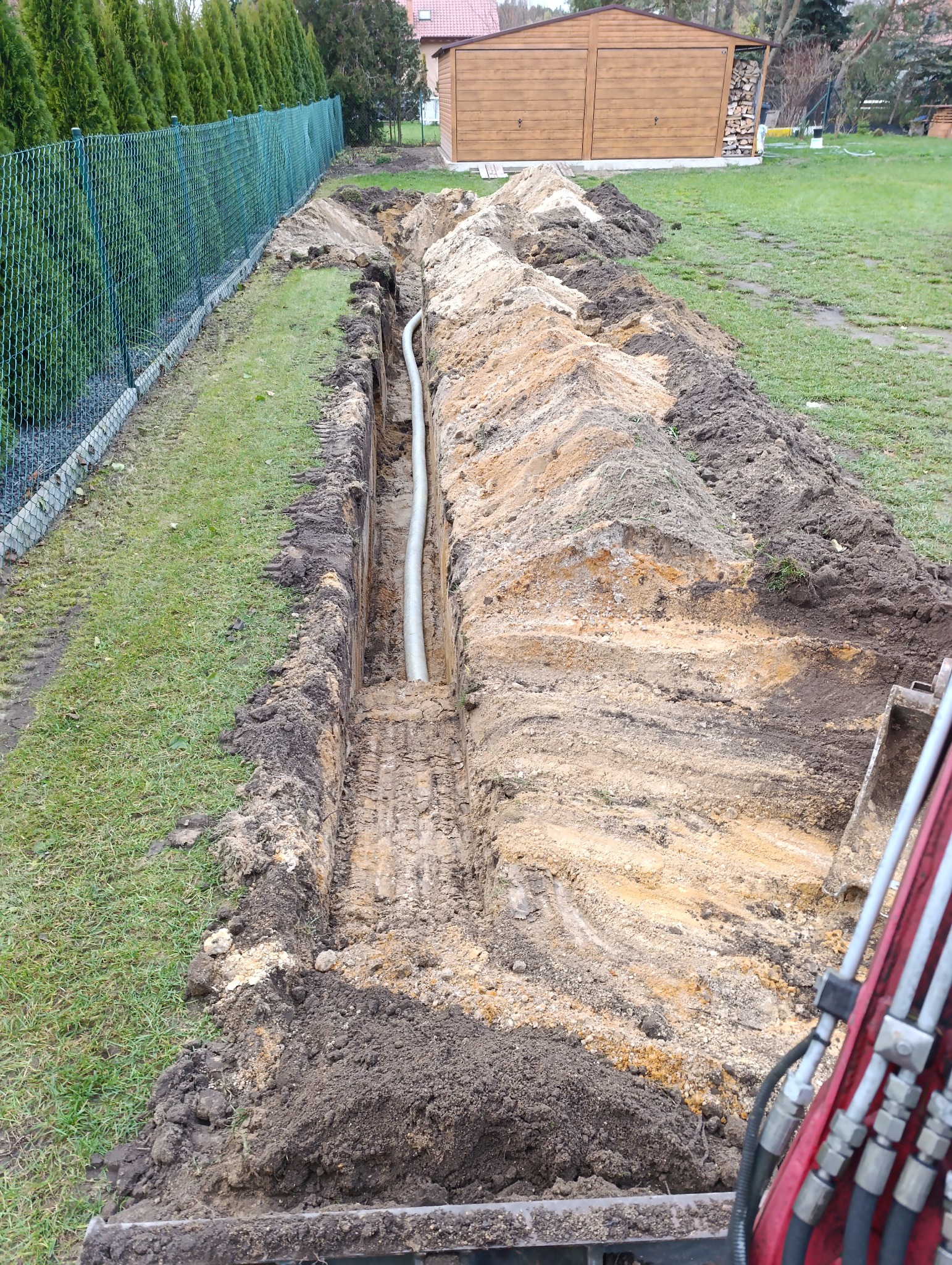 Wykopany rów w ziemi z ułożoną szarą rurą, widoczne ślady opon, zielony trawnik, ogrodzenie z siatki i budynek gospodarczy w tle.