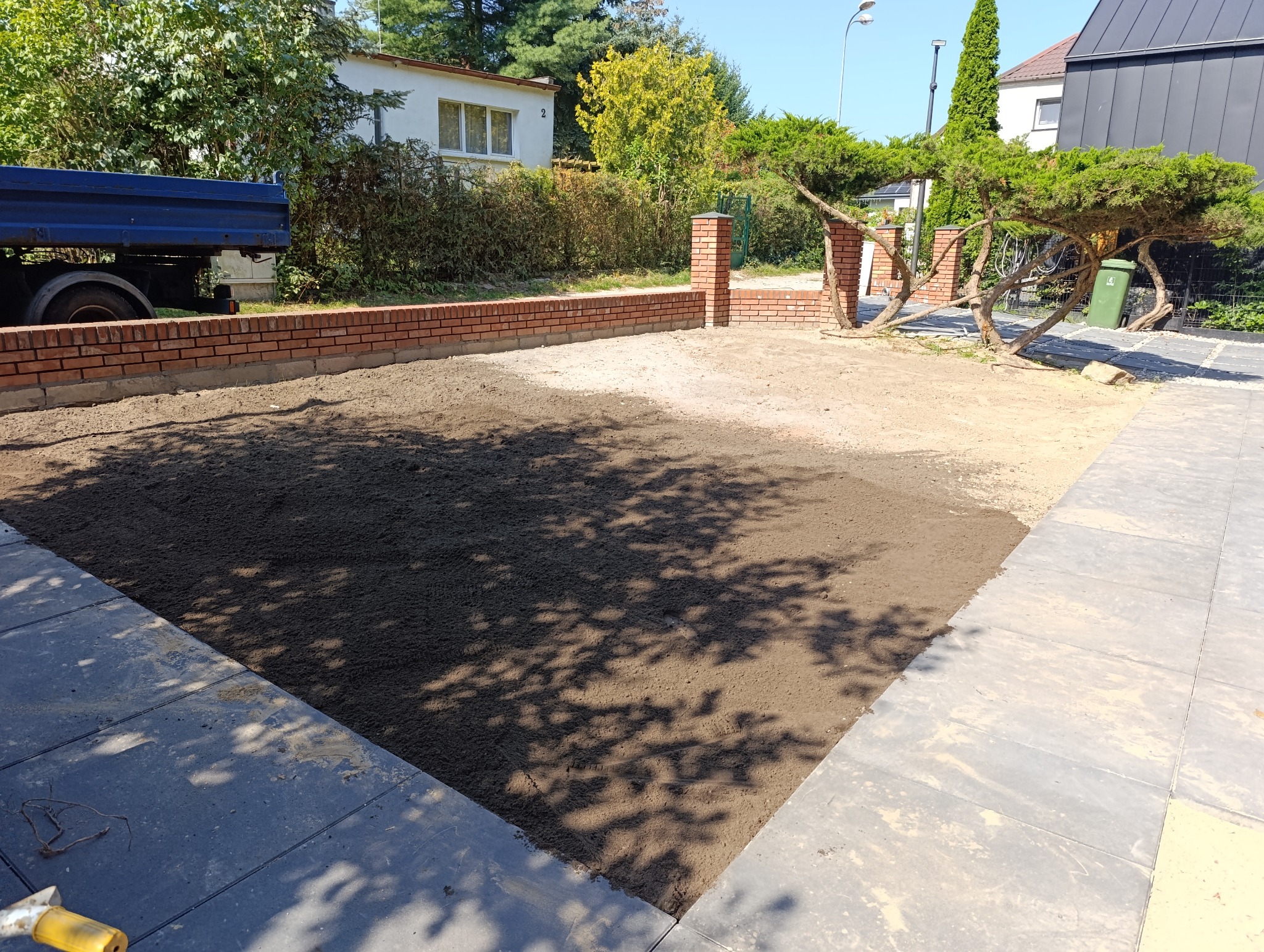 Przygotowany teren pod trawnik z widoczną świeżo ułożoną ziemią, obrzeżami z kostki i fragmentem murku z cegły. W tle budynki i zieleń.