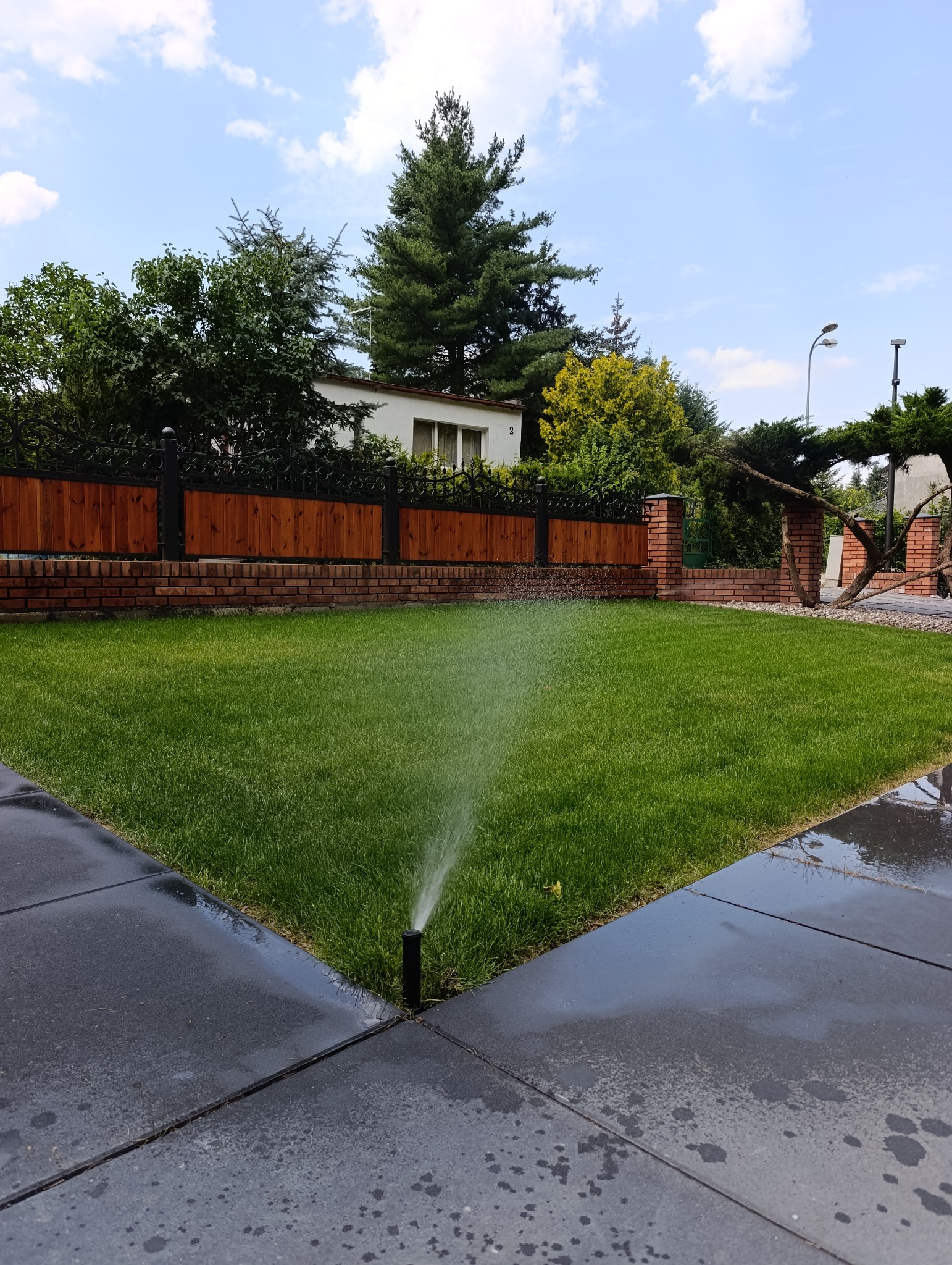 Automatyczny system nawadniania trawnika w ogrodzie. Zraszacze w akcji, nawadniające soczystą zieleń trawy. W tle ogrodzenie z drewnianymi elementami i ceglanymi słupkami.