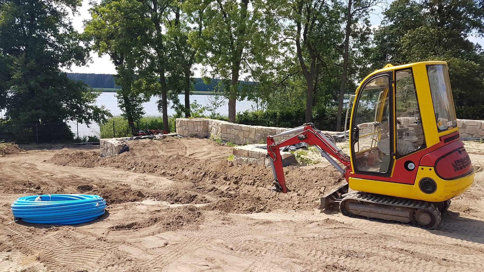 Żółto-czerwona mini koparka na placu budowy ogrodu z kamiennym murem w tle i zwiniętymi niebieskimi rurami. Jezioro i drzewa w oddali.