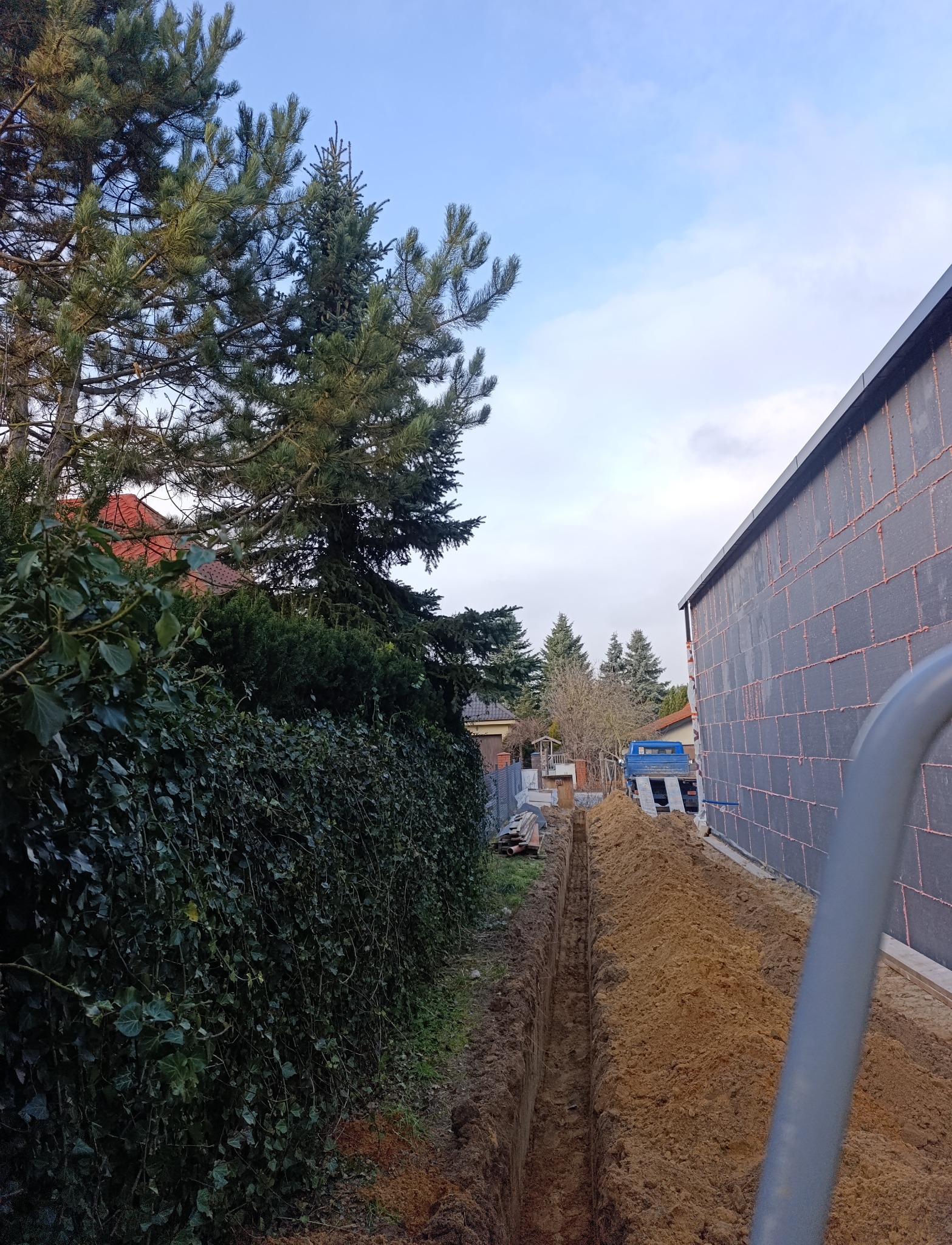 Wykop pod system nawadniania ogrodu, widoczny z perspektywy ziemi. W tle zieleń, budynek w trakcie budowy i błękitne niebo. Wykop równy, wąski, z wyraźnymi ścianami.