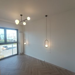 Roman Plikhivskyi - Pusty pokój z parkietem w jodełkę, nowoczesnym oświetleniem sufitowym i ściennym, białe ściany z dekoracyjnymi listwami, duże okno z widokiem na budynek mieszkalny.