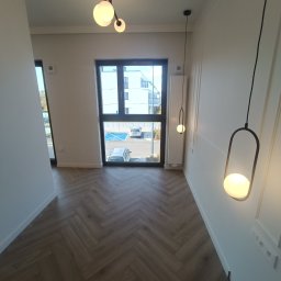 Roman Plikhivskyi - Wnętrze przedpokoju z parkietem ułożonym w jodełkę, wiszącymi lampami z okrągłymi kloszami i widokiem na parking przez okno.