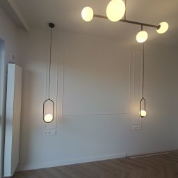 Roman Plikhivskyi - Nowoczesne wnętrze z białymi ścianami zdobionymi dekoracyjnymi listwami, parkietem w jodełkę i designerskim oświetleniem: lampa sufitowa z kilkoma kulistymi kloszami i dwie wiszące lampy z czarnymi...