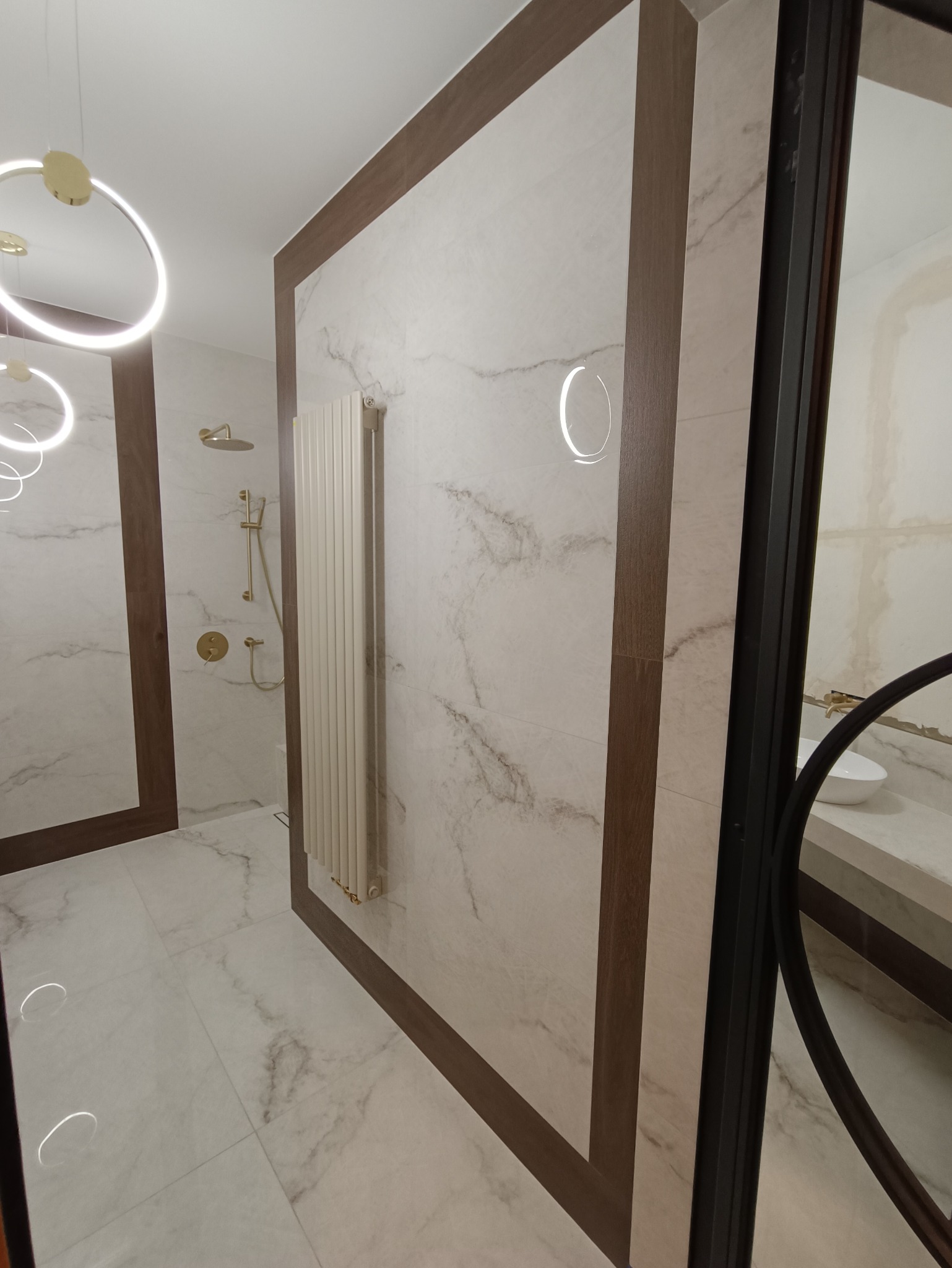 Nowoczesna łazienka z marmurowymi płytkami, grzejnikiem drabinkowym w kolorze kremowym, złotą armaturą prysznicową i okrągłymi lampami LED na suficie, widok przez częściowo zamontowaną czarną...
