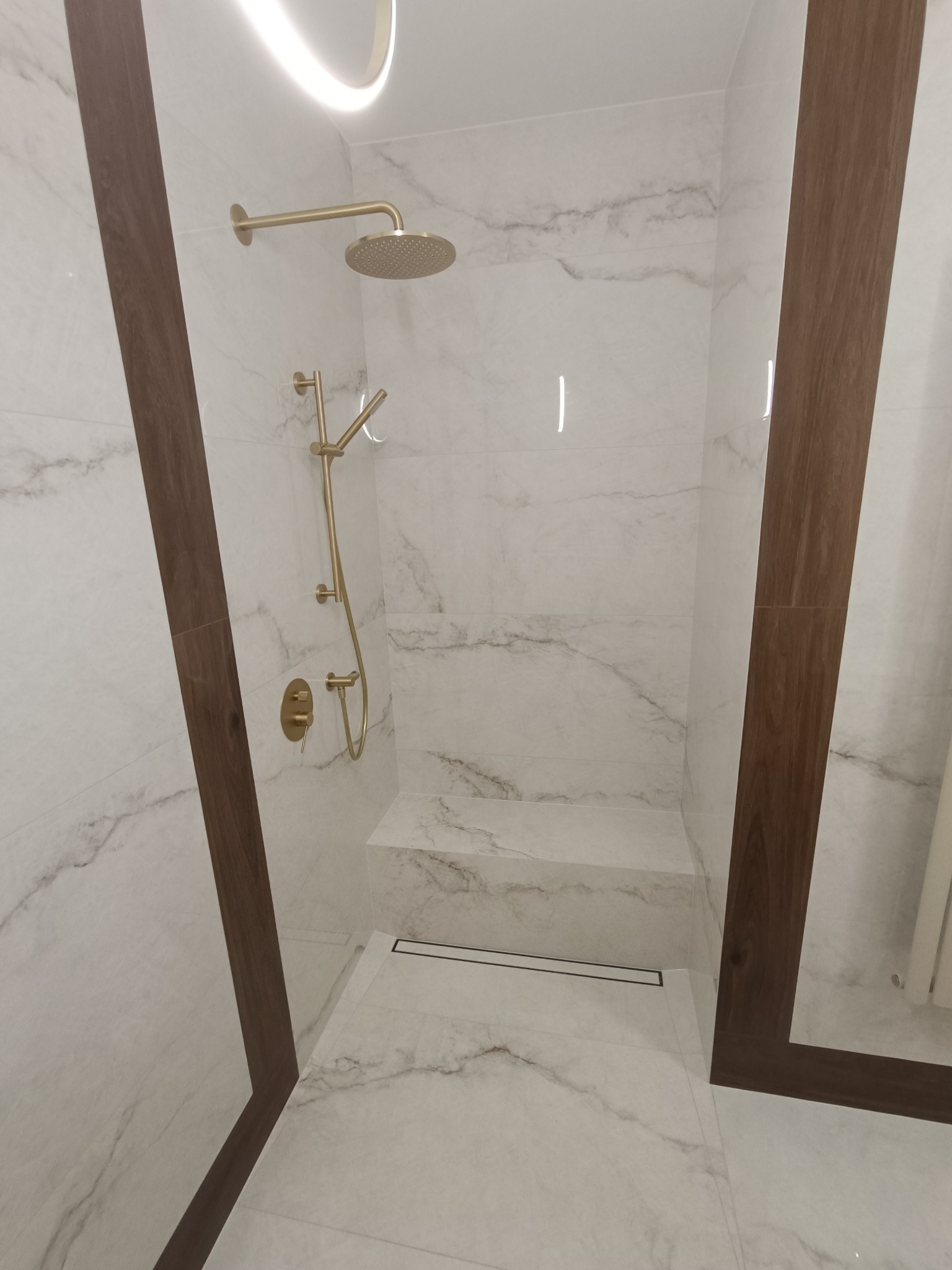 Nowoczesna kabina prysznicowa z marmurowymi płytkami, złotą armaturą i drewnianymi akcentami wykończeniowymi.
