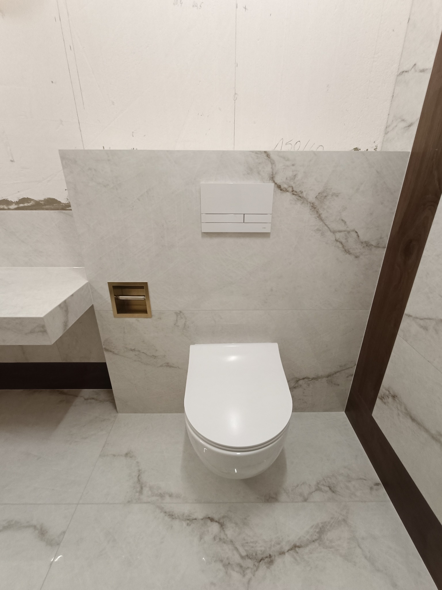 Nowoczesna łazienka w trakcie wykańczania: podwieszana toaleta, marmurowe płytki na ścianach i podłodze, zabudowana półka z umywalką, złoty pojemnik na papier toaletowy, fragment drewnianej ramy...