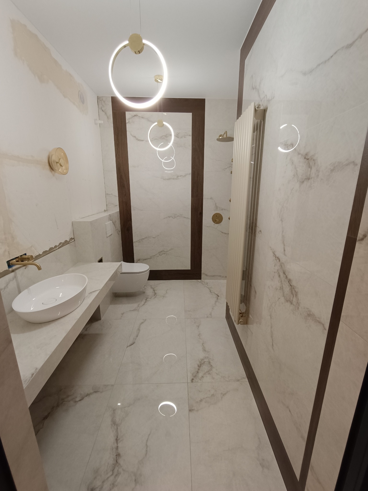 Wnętrze łazienki w trakcie prac wykończeniowych, widoczne marmurowe płytki, biała ceramika sanitarna, złota armatura i okrągłe lampy sufitowe, ściany częściowo przygotowane pod malowanie.