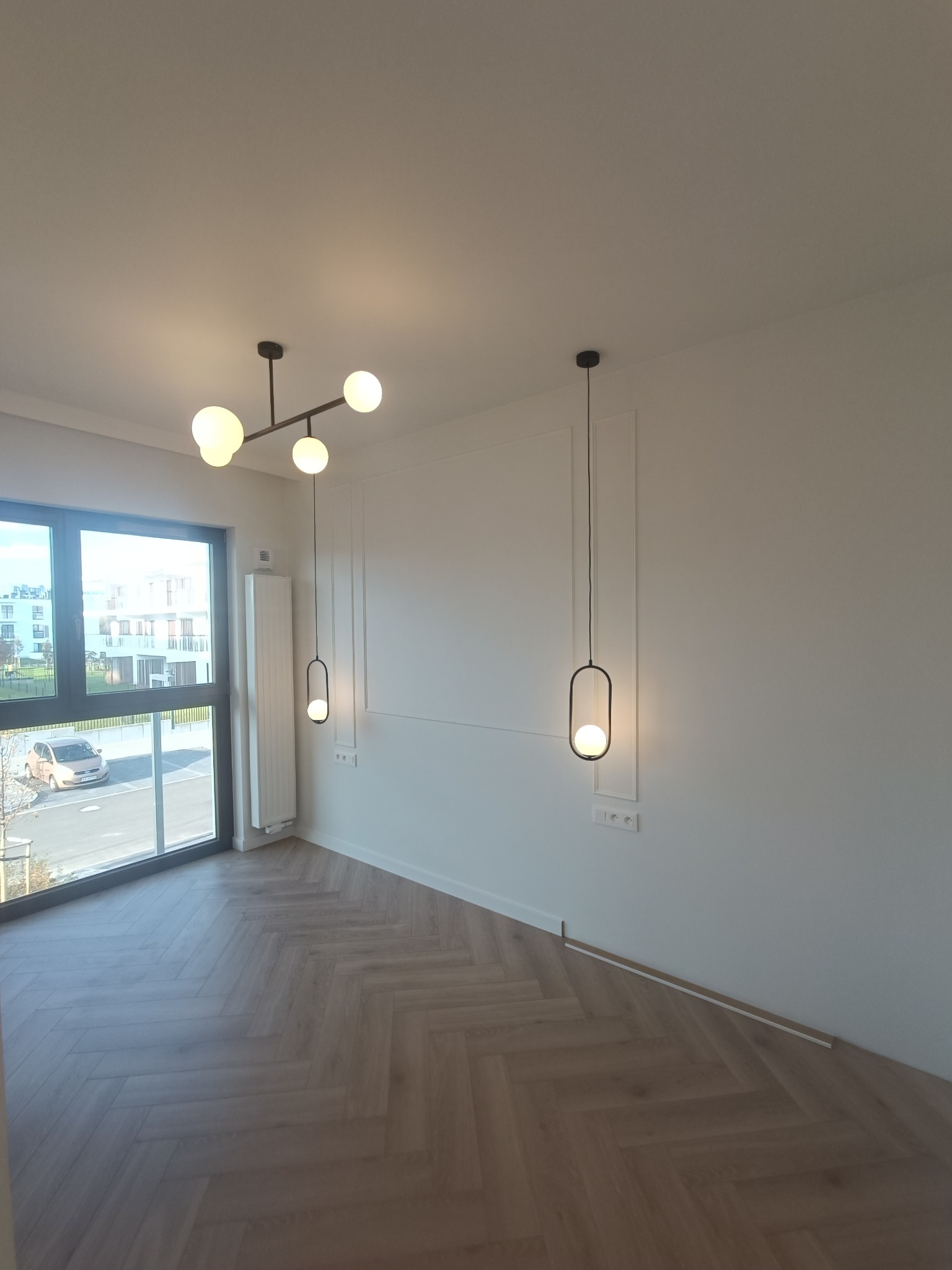 Pusty pokój z parkietem w jodełkę, nowoczesnym oświetleniem sufitowym i ściennym, białe ściany z dekoracyjnymi listwami, duże okno z widokiem na budynek mieszkalny.