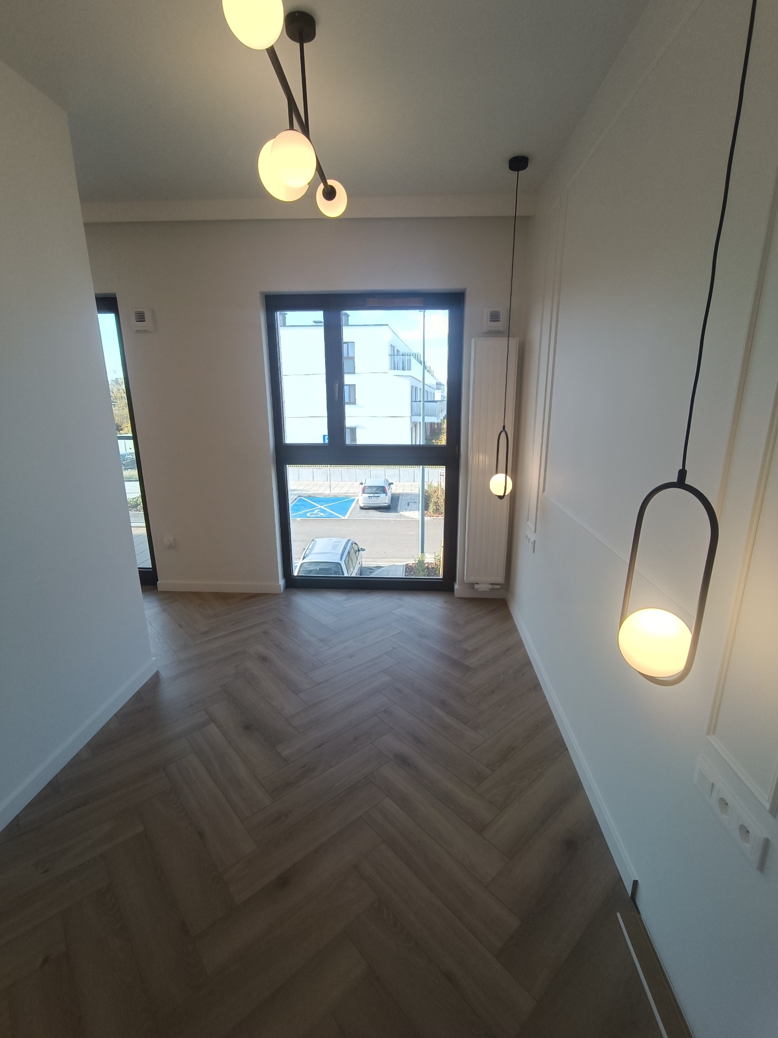 Wnętrze przedpokoju z parkietem ułożonym w jodełkę, wiszącymi lampami z okrągłymi kloszami i widokiem na parking przez okno.