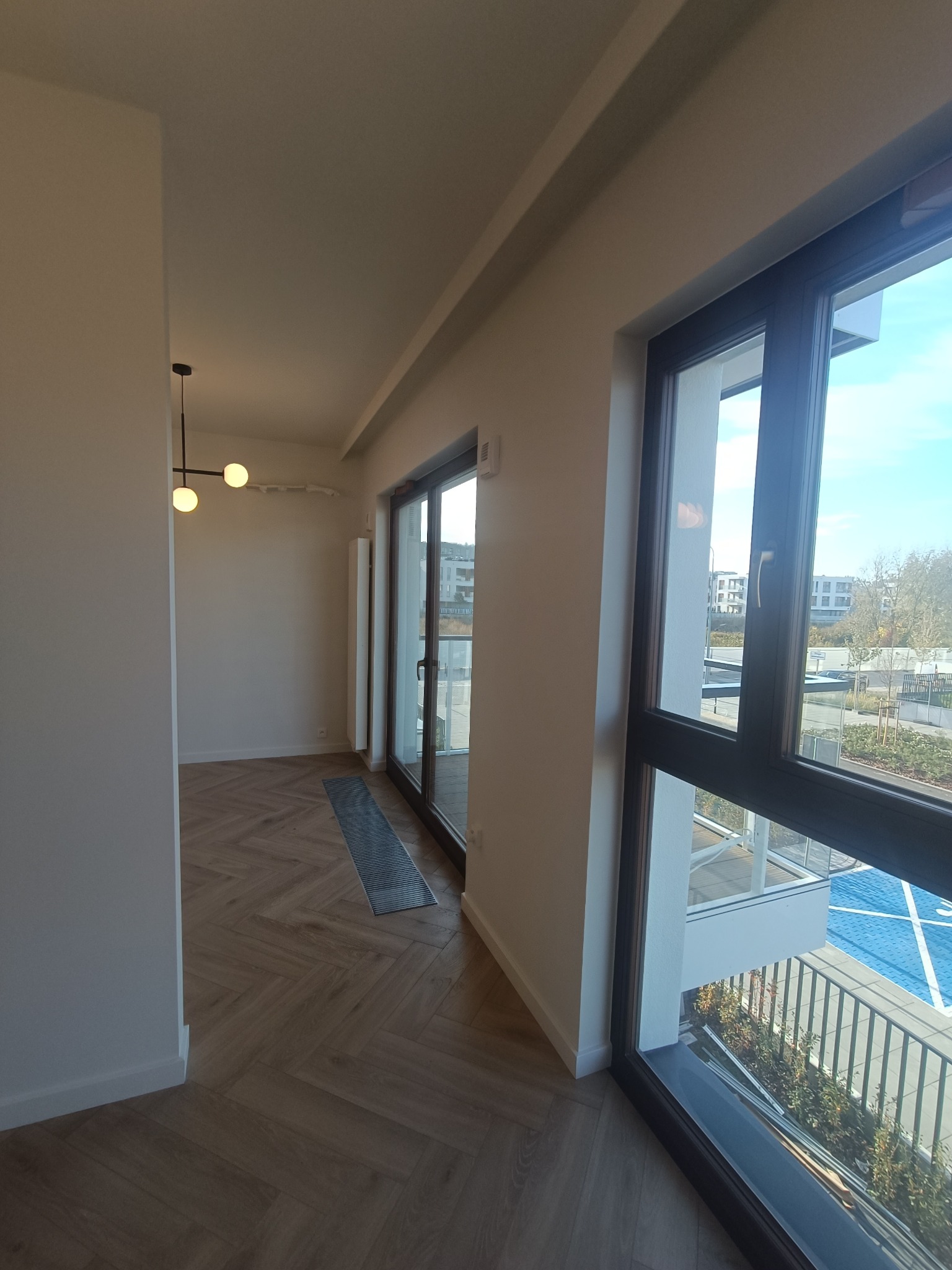 Wnętrze mieszkania po remoncie: parkiet ułożony w jodełkę, duże okna balkonowe z widokiem na zewnątrz, nowoczesna lampa sufitowa.