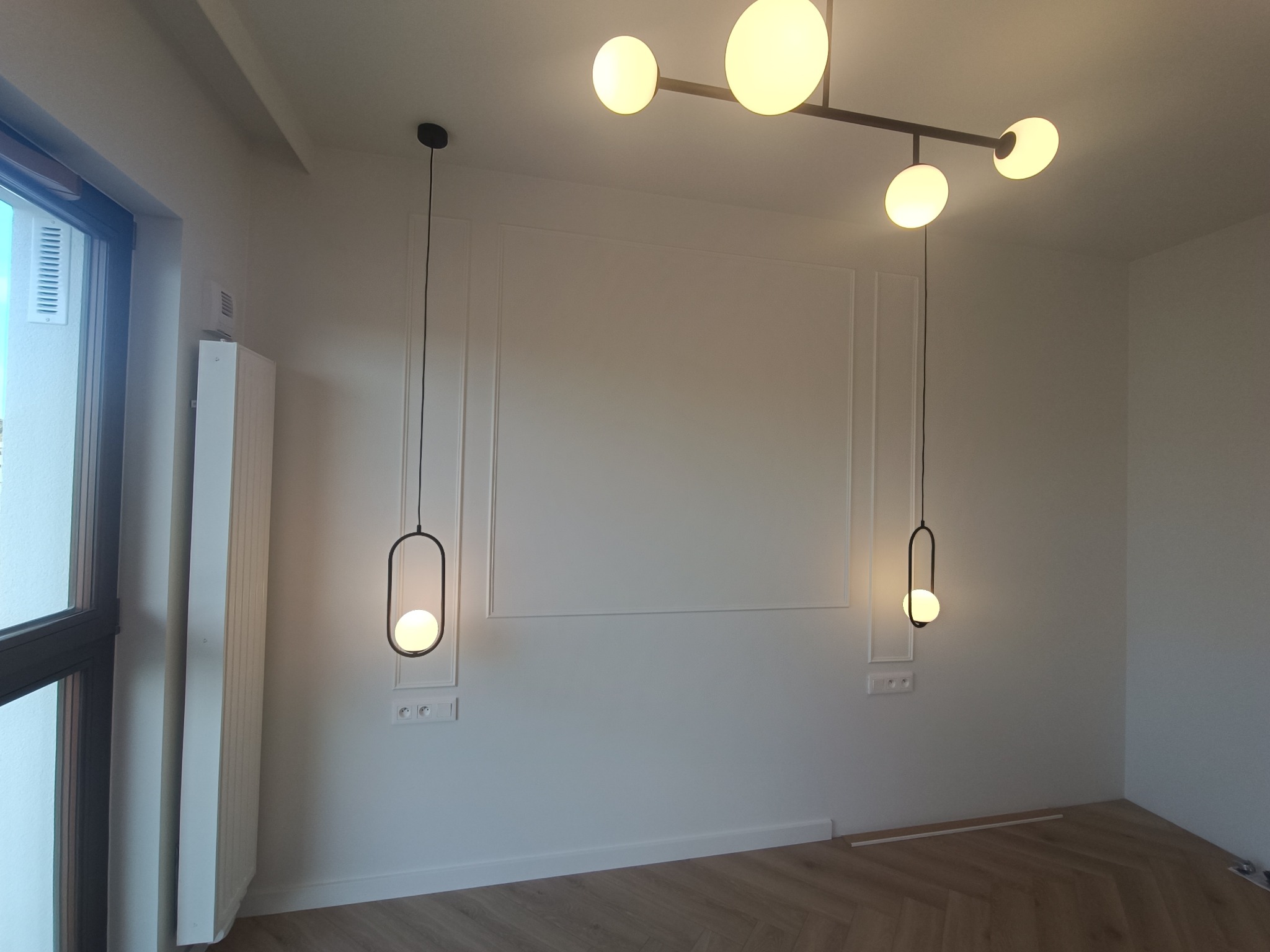 Nowoczesne wnętrze z białymi ścianami zdobionymi dekoracyjnymi listwami, parkietem w jodełkę i designerskim oświetleniem: lampa sufitowa z kilkoma kulistymi kloszami i dwie wiszące lampy z czarnymi...