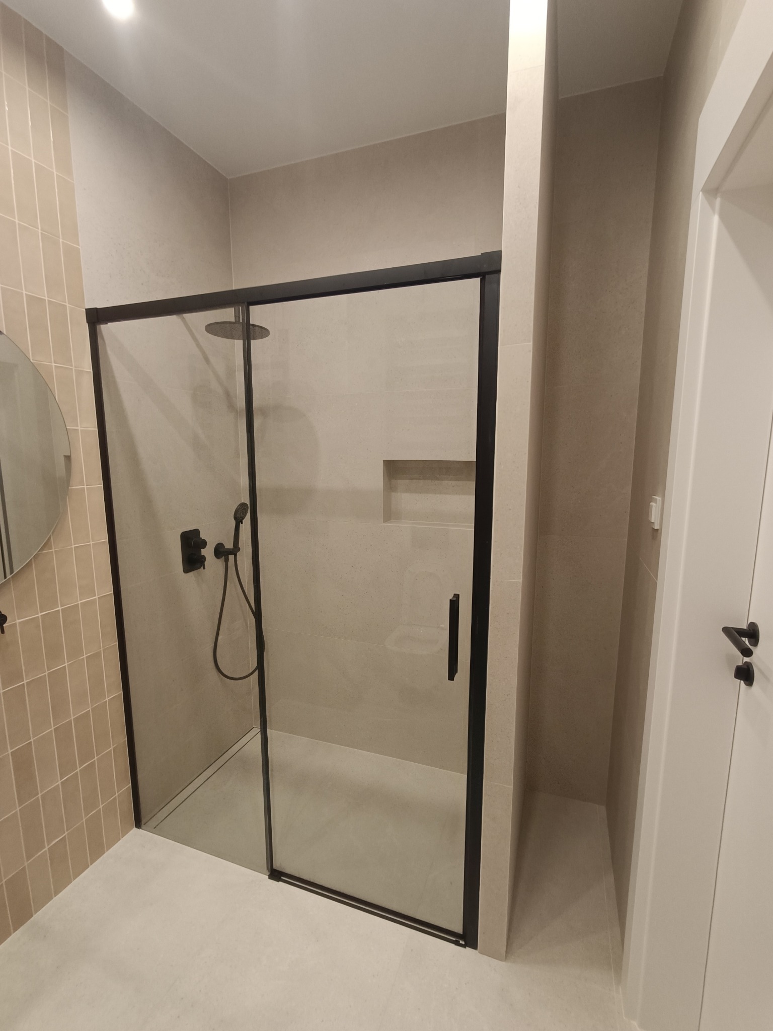 Nowoczesna kabina prysznicowa z czarnymi profilami i deszczownicą, beżowe płytki na ścianach i podłodze, okrągłe lustro po lewej stronie, widok na fragment białych drzwi po prawej.