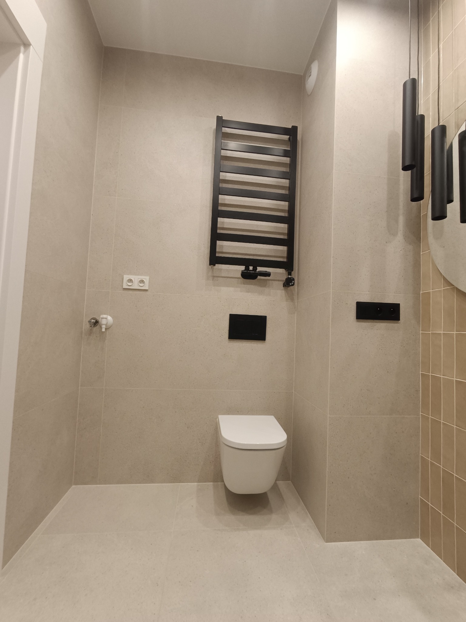 Nowoczesna łazienka z beżowymi płytkami, wiszącą toaletą, grzejnikiem drabinkowym i czarnymi akcentami, w tym włącznikami i lampą wiszącą, z widocznym fragmentem okrągłego lustra.