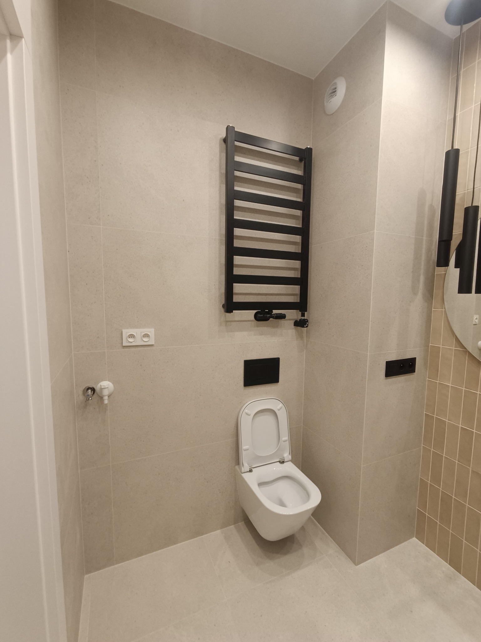 Nowoczesna łazienka wykończona w beżowych płytkach z podwieszaną toaletą, grzejnikiem drabinkowym w kolorze czarnym oraz okrągłym lustrem z czarnymi lampami.