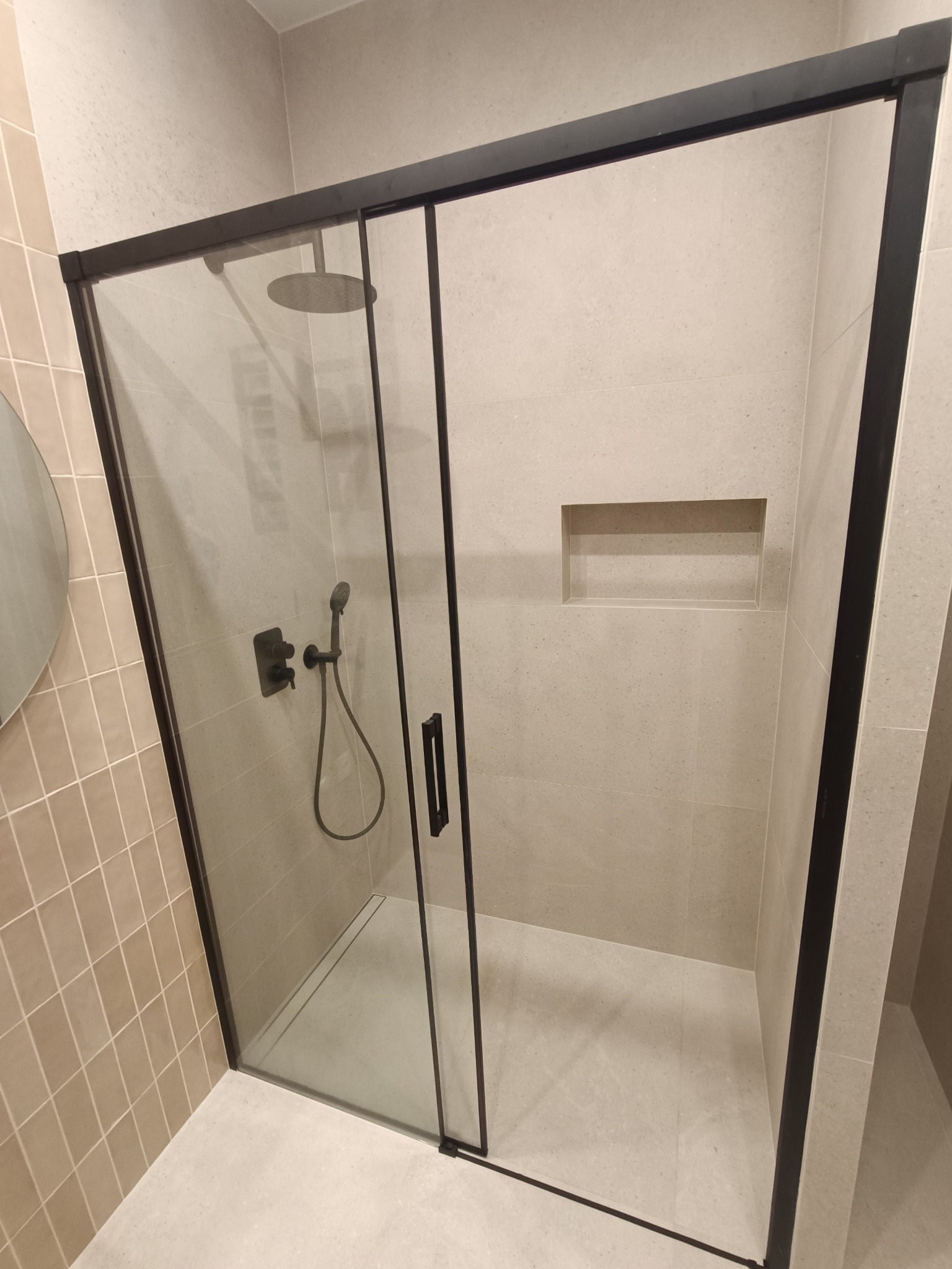 Nowoczesna kabina prysznicowa z czarnymi profilami i deszczownicą, w łazience wykończonej jasnymi płytkami z wnęką.