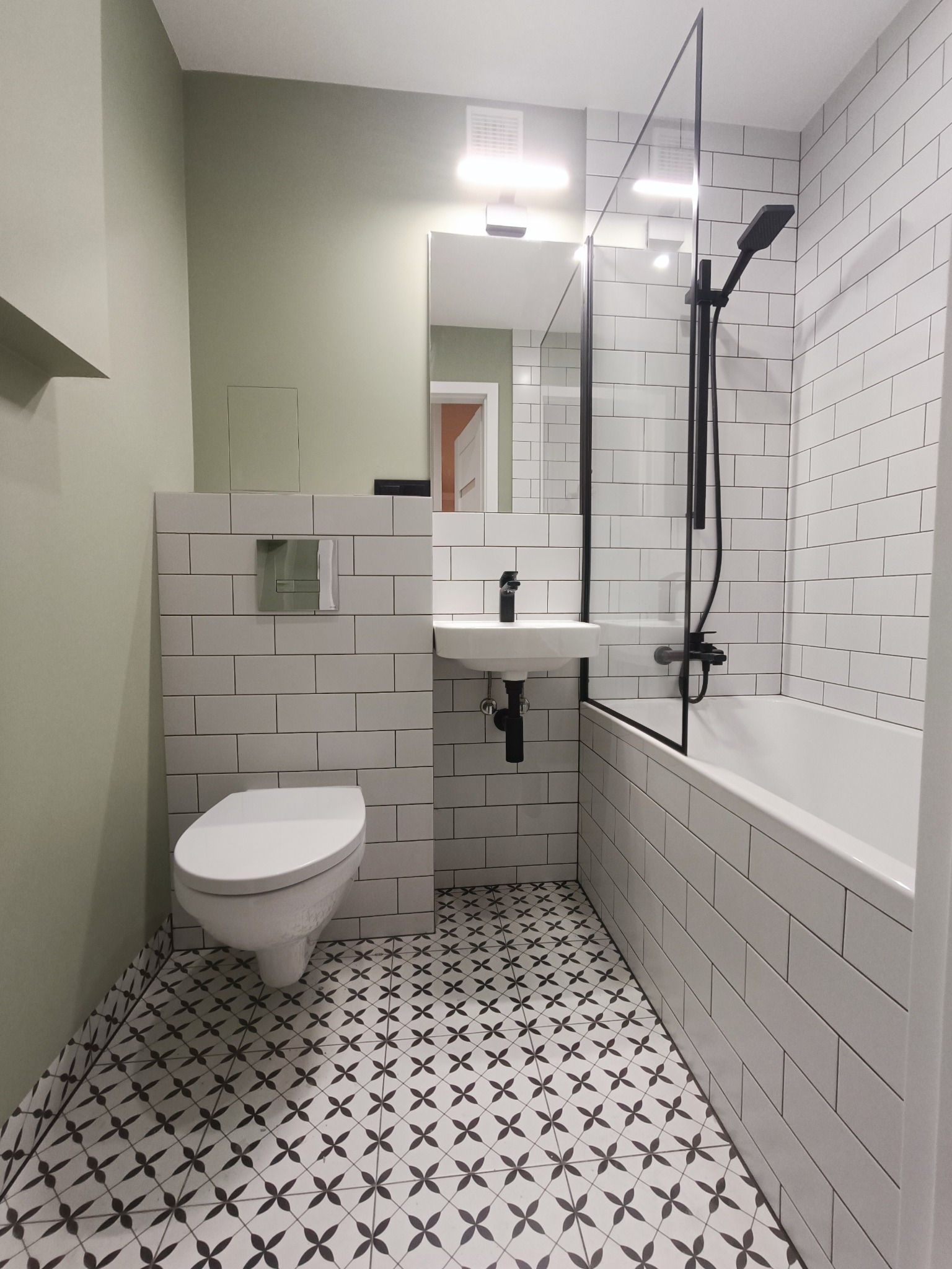 Wykończona łazienka z białymi płytkami ułożonymi w cegiełkę, czarną armaturą, toaletą podwieszaną i wzorzystą podłogą w stylu retro.