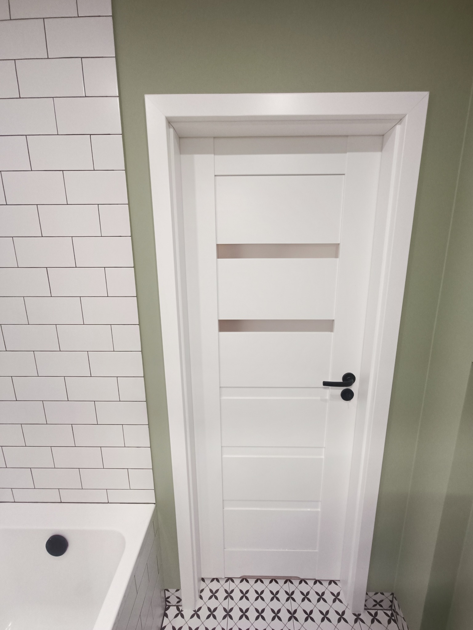 Biała, nowoczesna łazienka z drzwiami z poziomymi przeszkleniami, białe płytki na ścianie, wzorzysta podłoga i oliwkowa ściana.