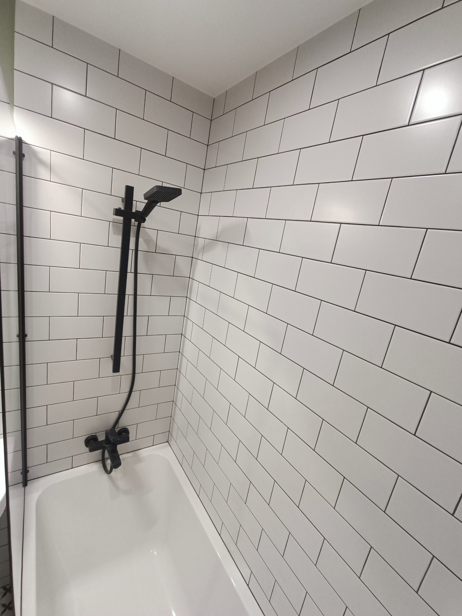Białe, prostokątne płytki ułożone w rzędach na ścianie nad wanną z czarną armaturą natryskową.