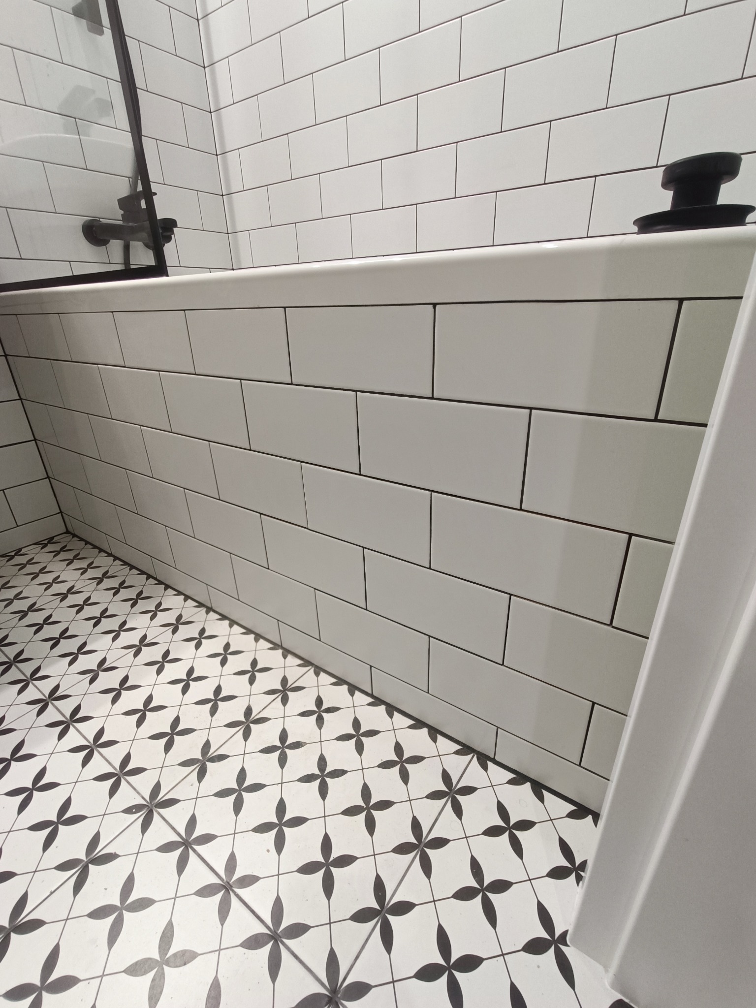 Białe, prostokątne płytki ścienne z ciemną fugą w łazience, kontrastujące z czarno-białą, wzorzystą podłogą z motywem kwiatowym, widoczny fragment czarnej armatury i białej wanny.