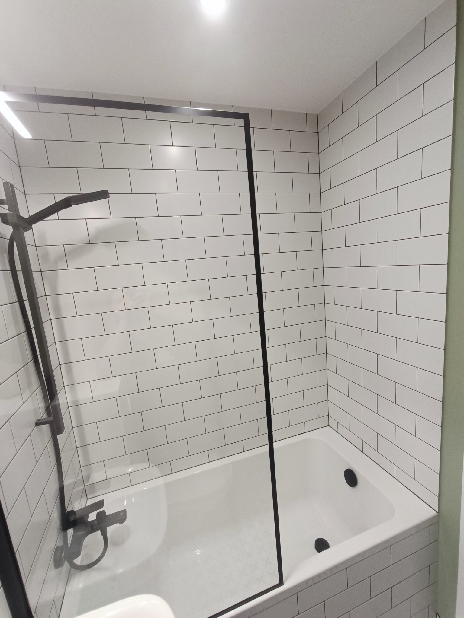 Biała łazienka z wanną obłożoną białymi, prostokątnymi płytkami, czarna armatura i przeszklona kabina prysznicowa z czarnymi ramami.