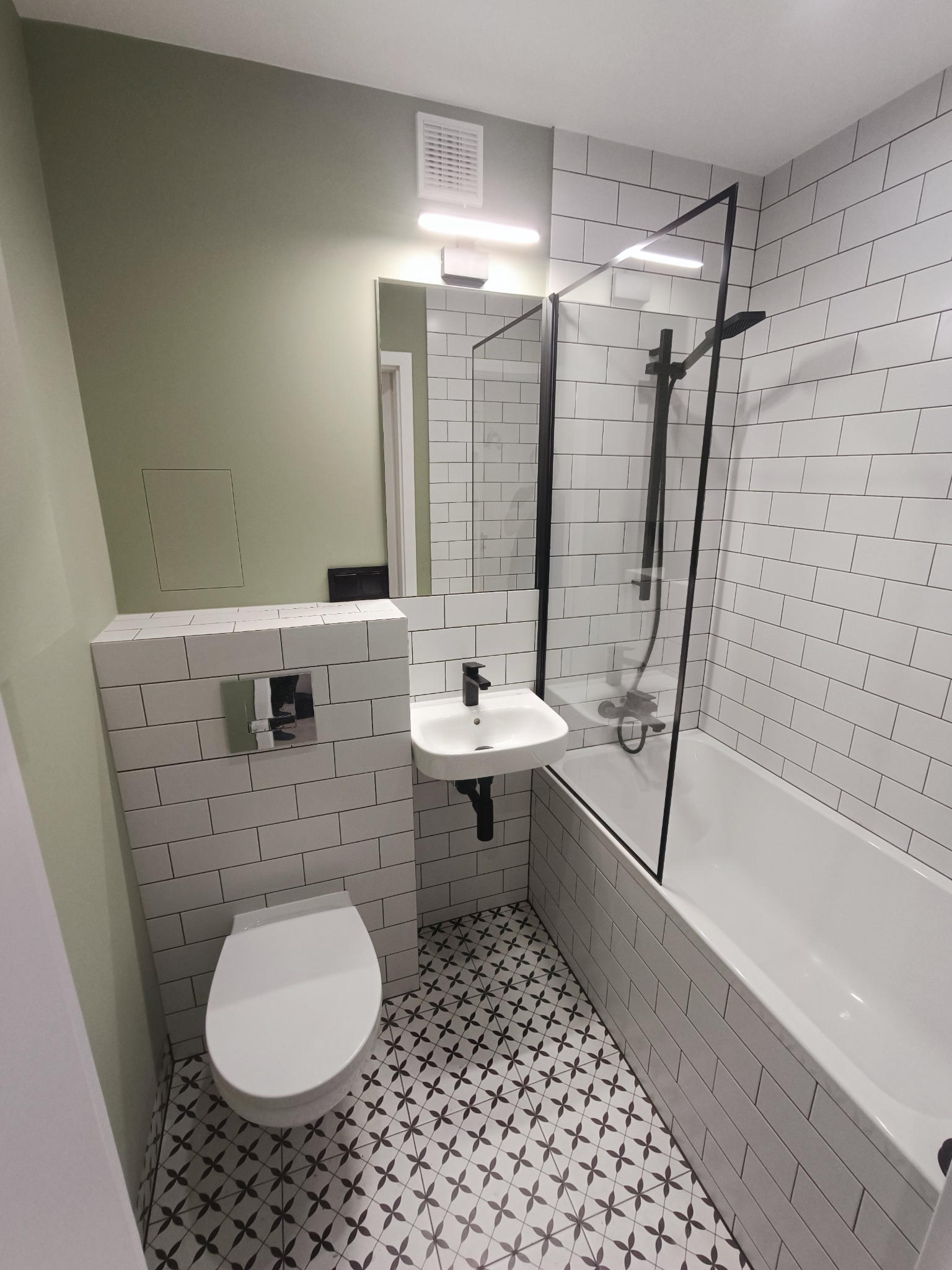Nowoczesna łazienka po remoncie: białe płytki imitujące cegłę, wzorzysta podłoga, minimalistyczna umywalka i czarna rama kabiny prysznicowej.