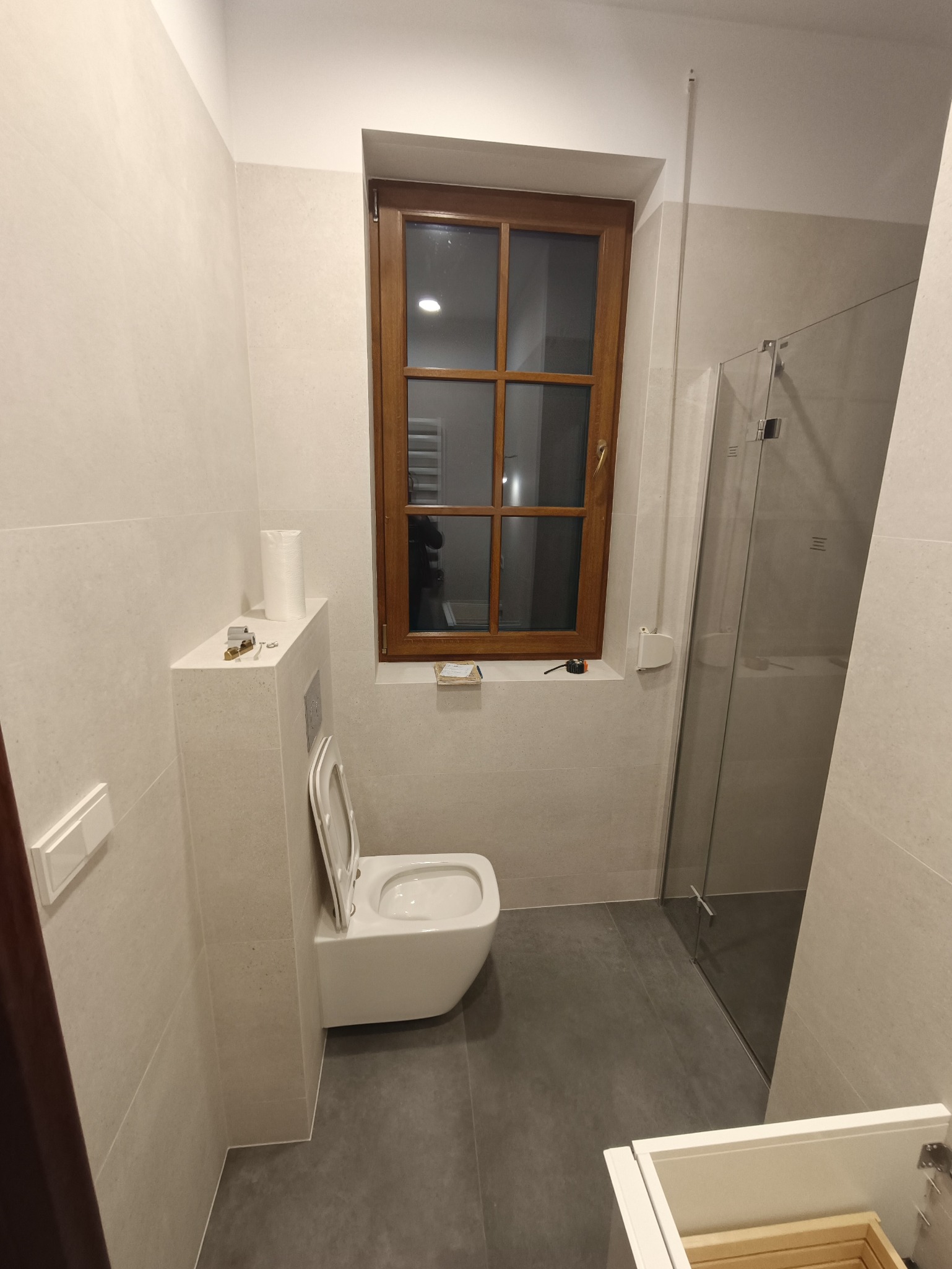 Wykończona łazienka z podwieszaną toaletą, szarą podłogą, kabiną prysznicową i drewnianym oknem, widoczne narzędzia i notatki na parapecie.