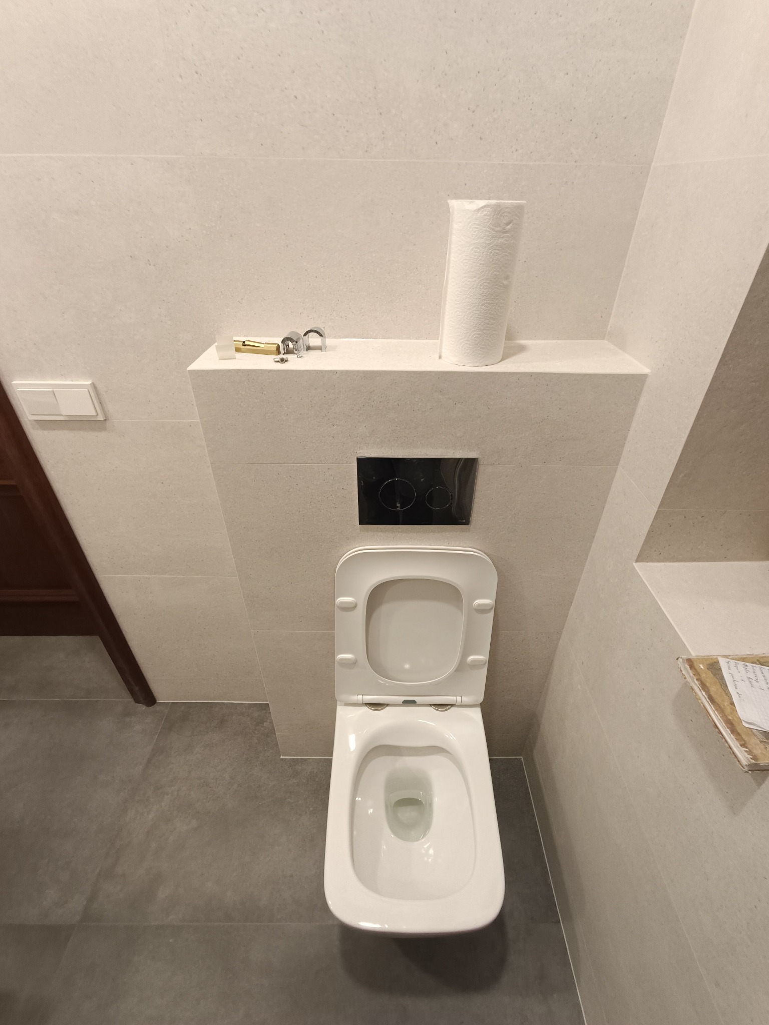 Nowoczesna toaleta podwieszana z otwartą klapą, szarymi płytkami podłogowymi i beżowymi płytkami ściennymi, rolka papieru toaletowego i elementy instalacyjne na półce, czarny przycisk spłuczki.