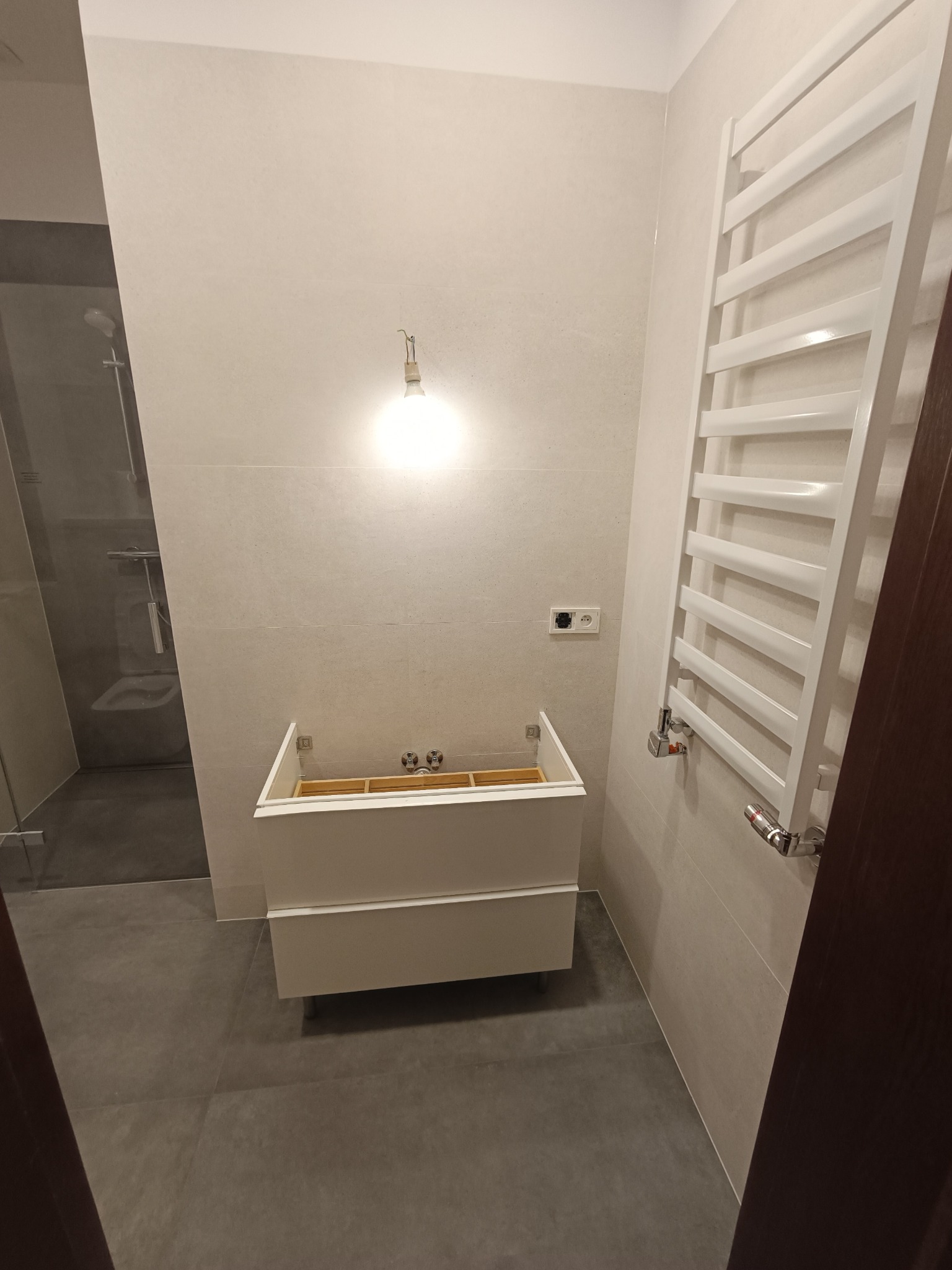 Niedokończona łazienka z zamontowaną białą szafką pod umywalkę bez blatu i baterii, grzejnikiem drabinkowym oraz widoczną kabiną prysznicową w tle, oświetlona pojedynczą żarówką.