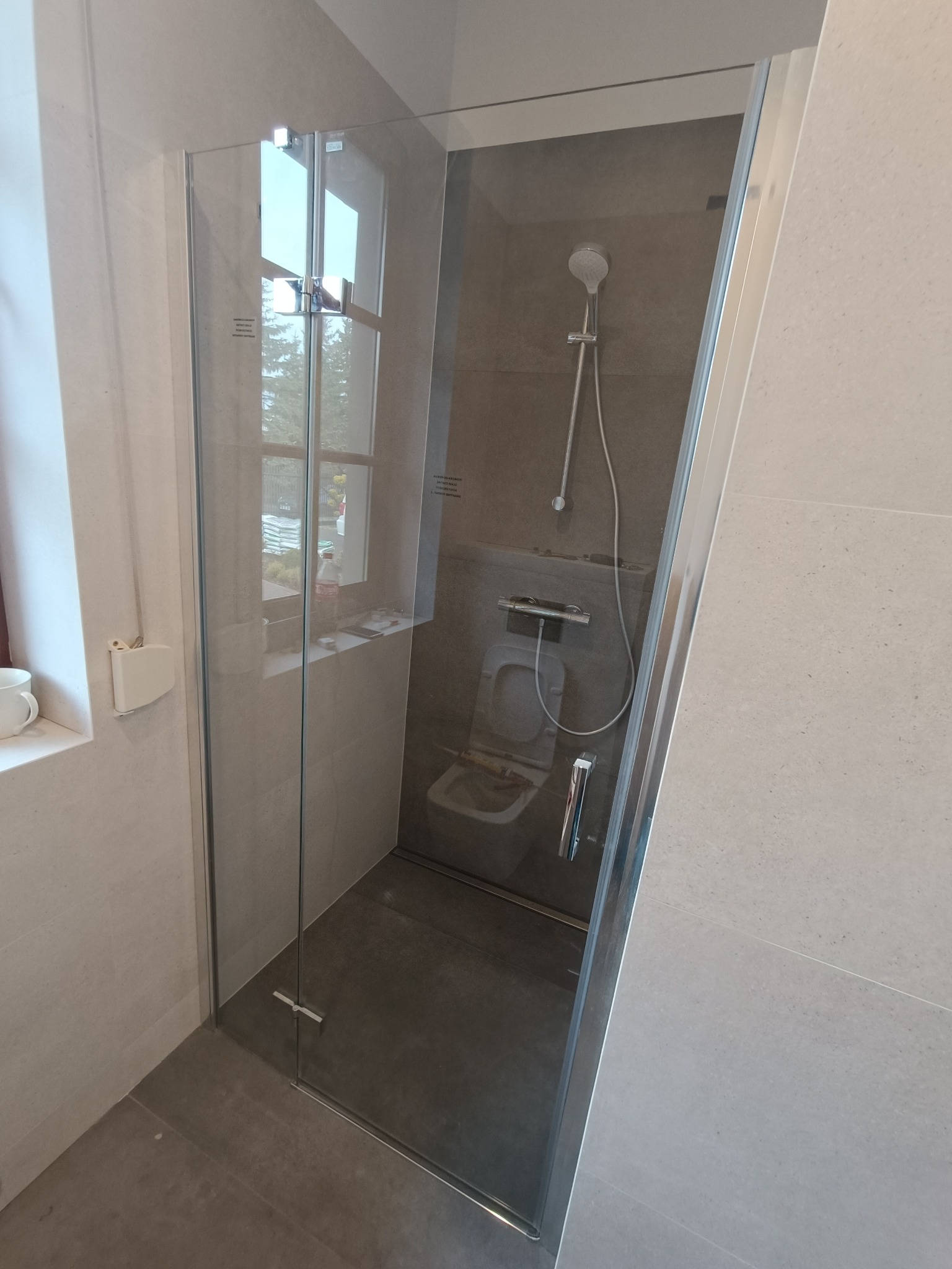 Kabina prysznicowa ze szklanymi drzwiami w nowoczesnej łazience w Warszawie, widoczny sedes podtynkowy i szara glazura.