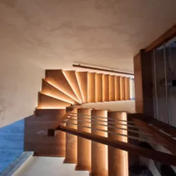 Kaskadowe schody z podświetleniem LED na stopniach, widziane z góry, z drewnianą balustradą o metalowych prętach. Neutralne tło ścian i sufitu.