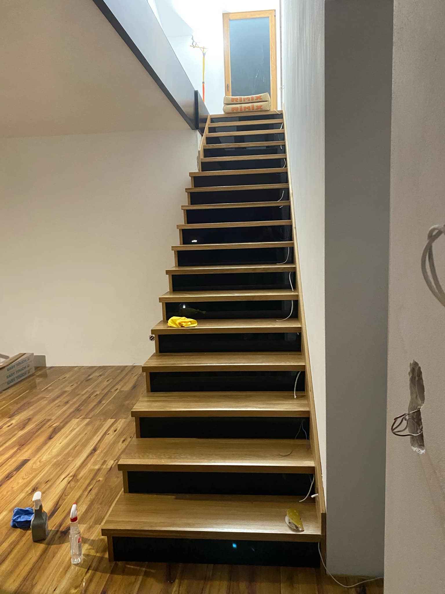 Nowoczesne schody drewniane z czarnymi podstopnicami w jasnym wnętrzu. Widoczne detale wykończenia i elementy przygotowania do dalszych prac.