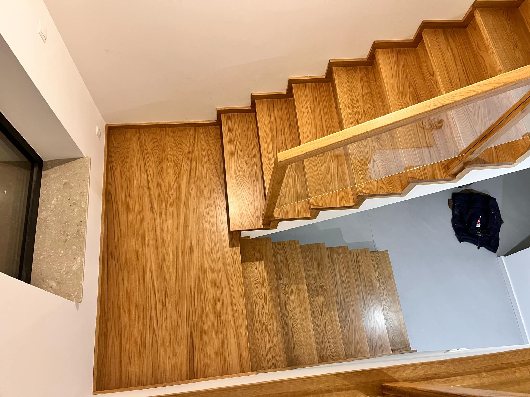 Widok z góry na nowoczesne schody z jasnego drewna z szklaną balustradą, z widocznym fragmentem okna i ściany.