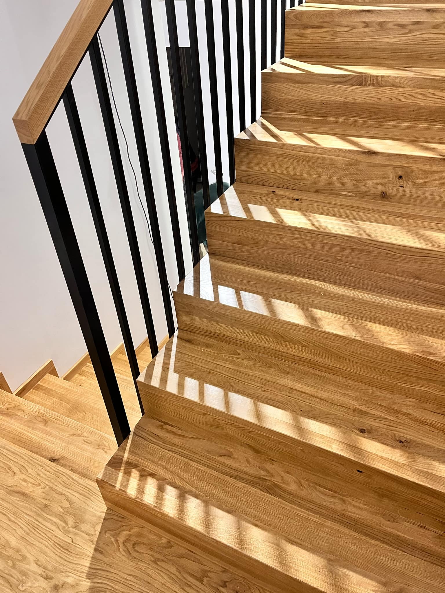 Jasne, dębowe schody z czarną, nowoczesną balustradą, oświetlone naturalnym światłem tworzącym cienie. Widok z góry na stopnie i fragment ściany.