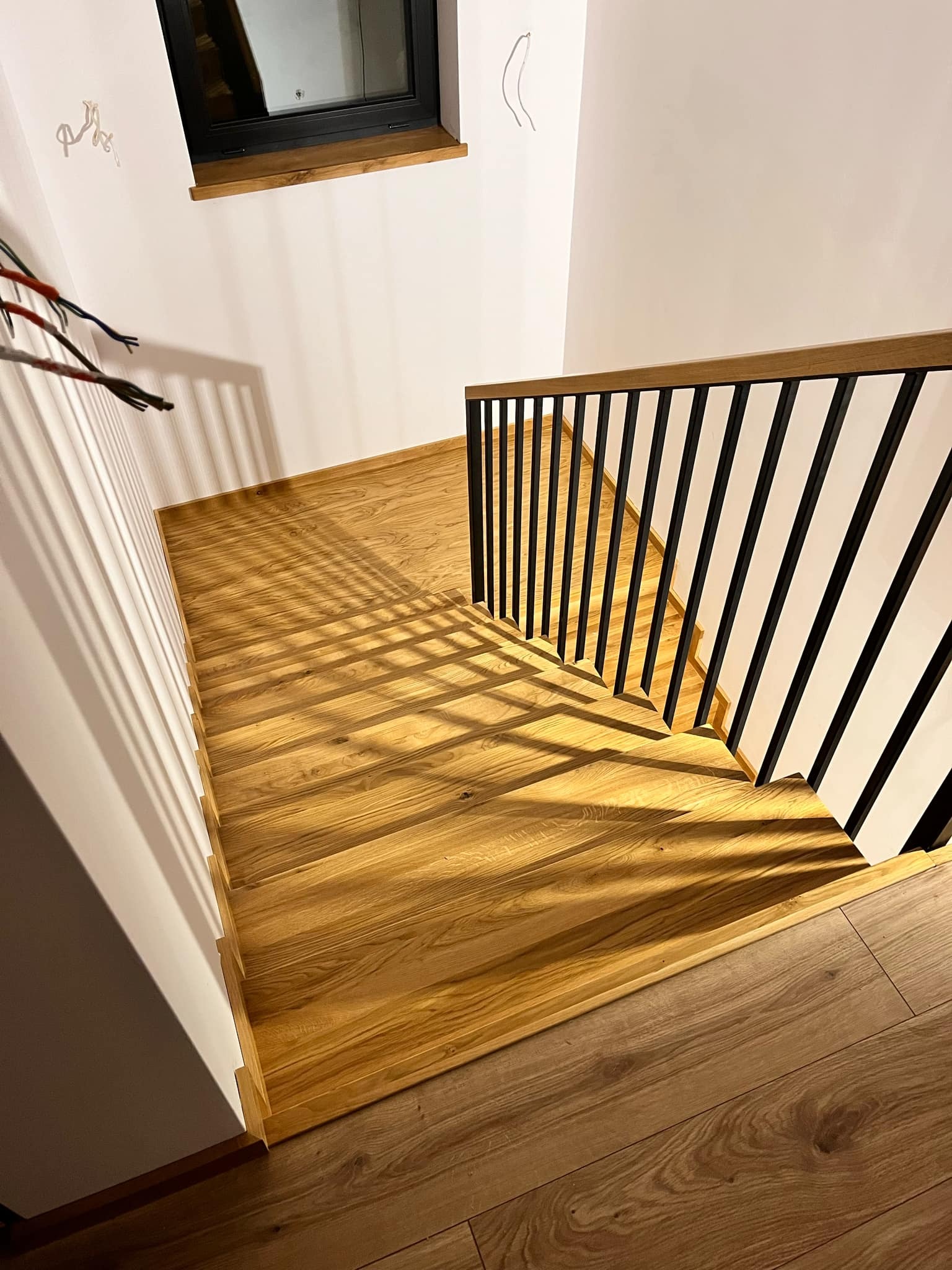 Nowoczesne schody z jasnego drewna z czarną metalową balustradą. Widok z góry, oświetlenie tworzy geometryczne cienie. U dołu widoczny fragment podłogi.